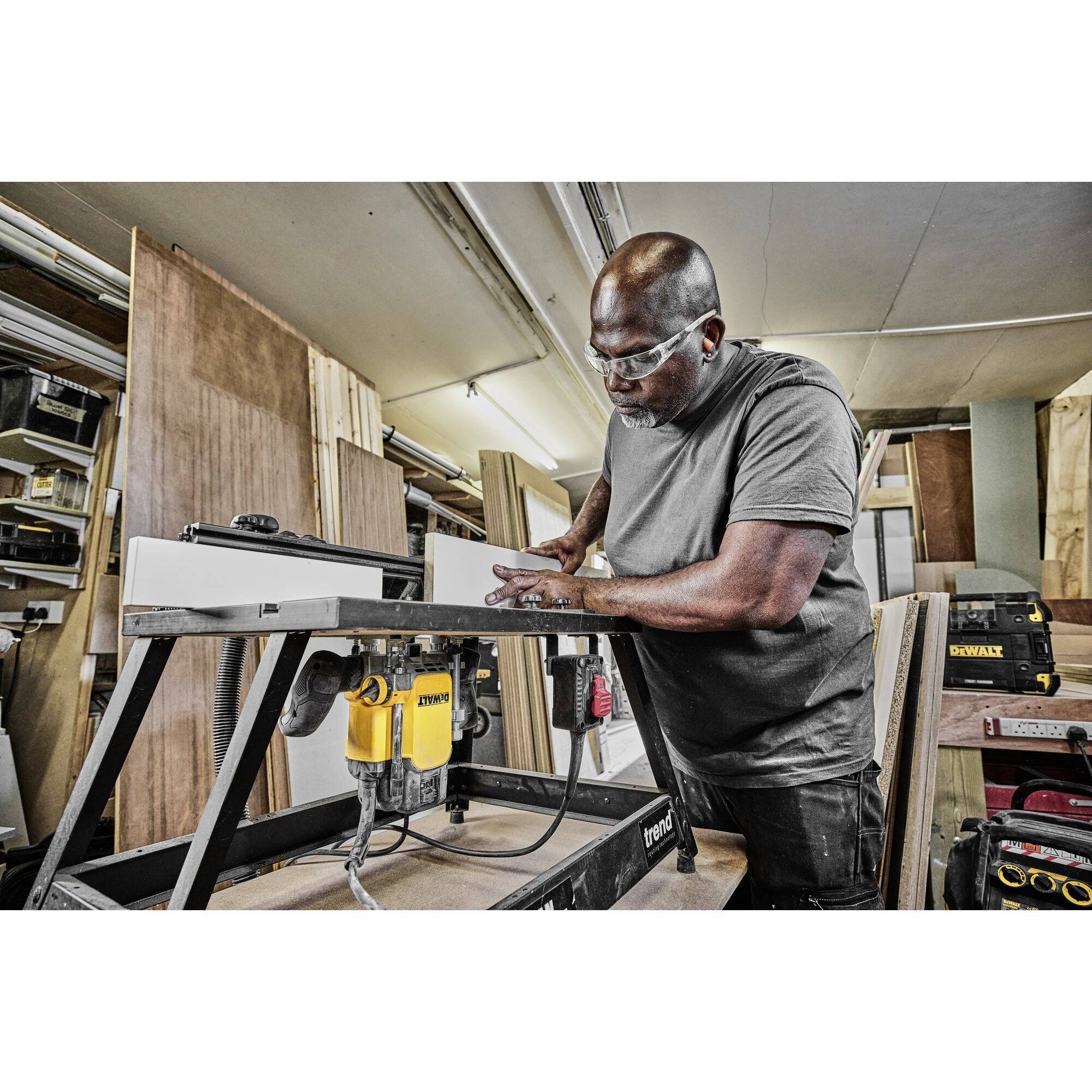 A man is working on a milling machine in a workshop, wearing safety glasses, surrounded by wooden materials and tools.