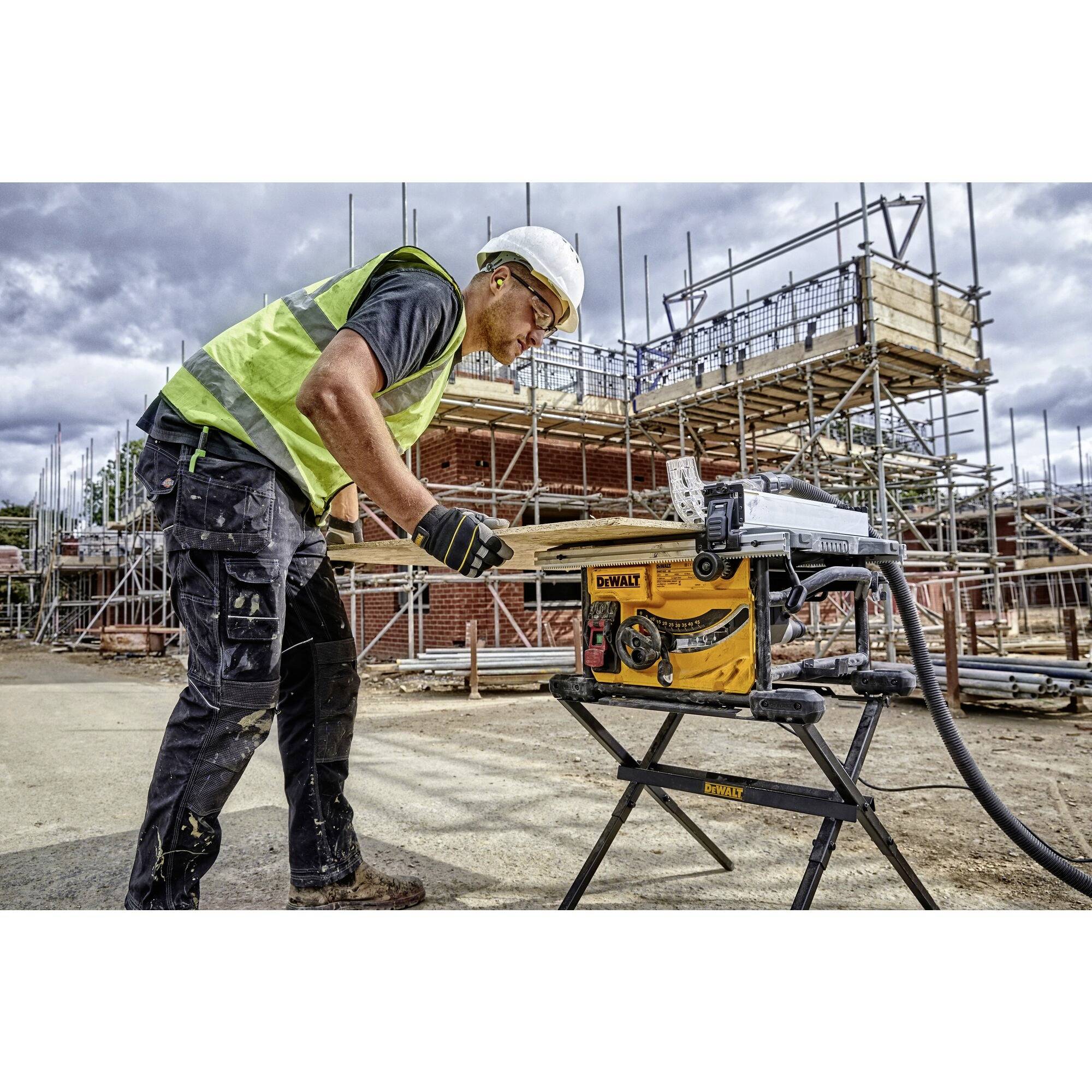 A construction worker wearing a high-visibility safety vest and hard hat is operating a table saw on a building site. Scaffolding can be seen in the background.