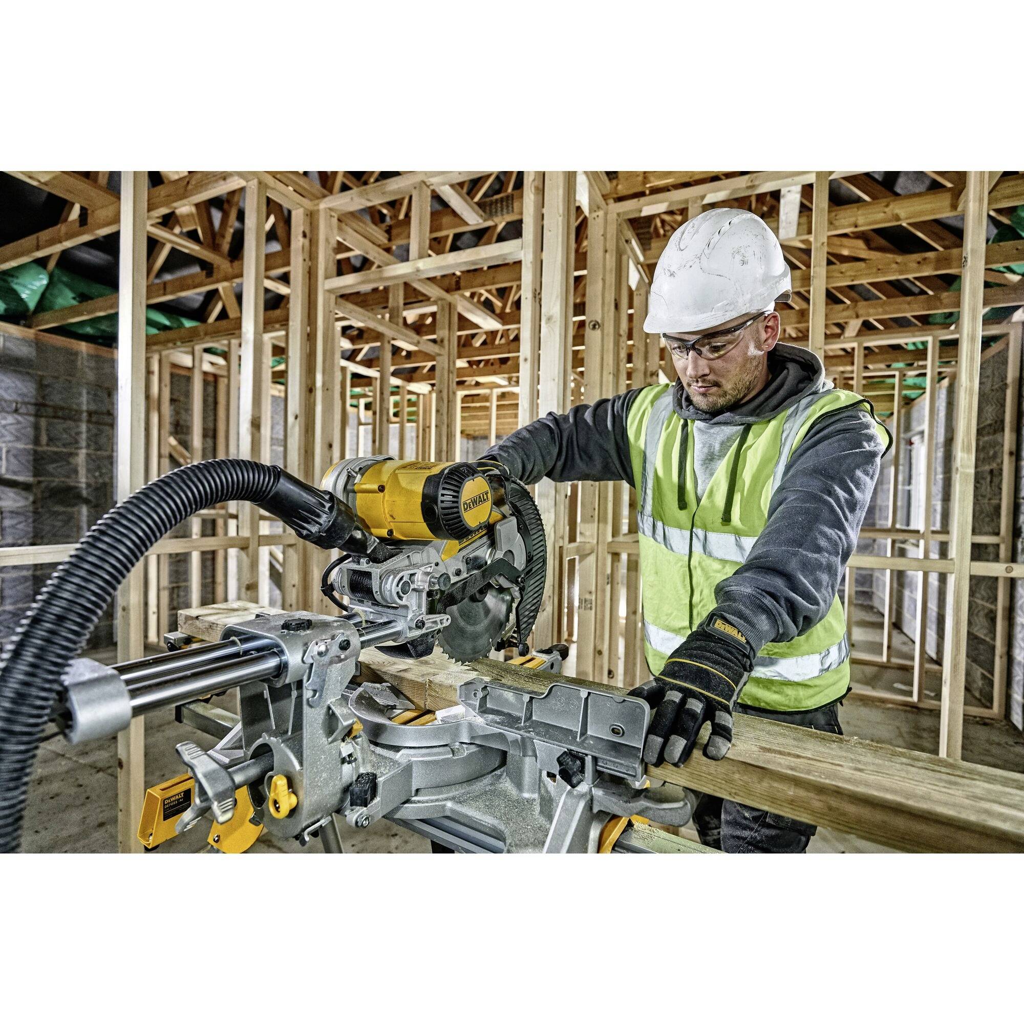 A construction worker wearing a hard hat and hi-vis vest is using an electric mitre saw to cut wood on a building site.
