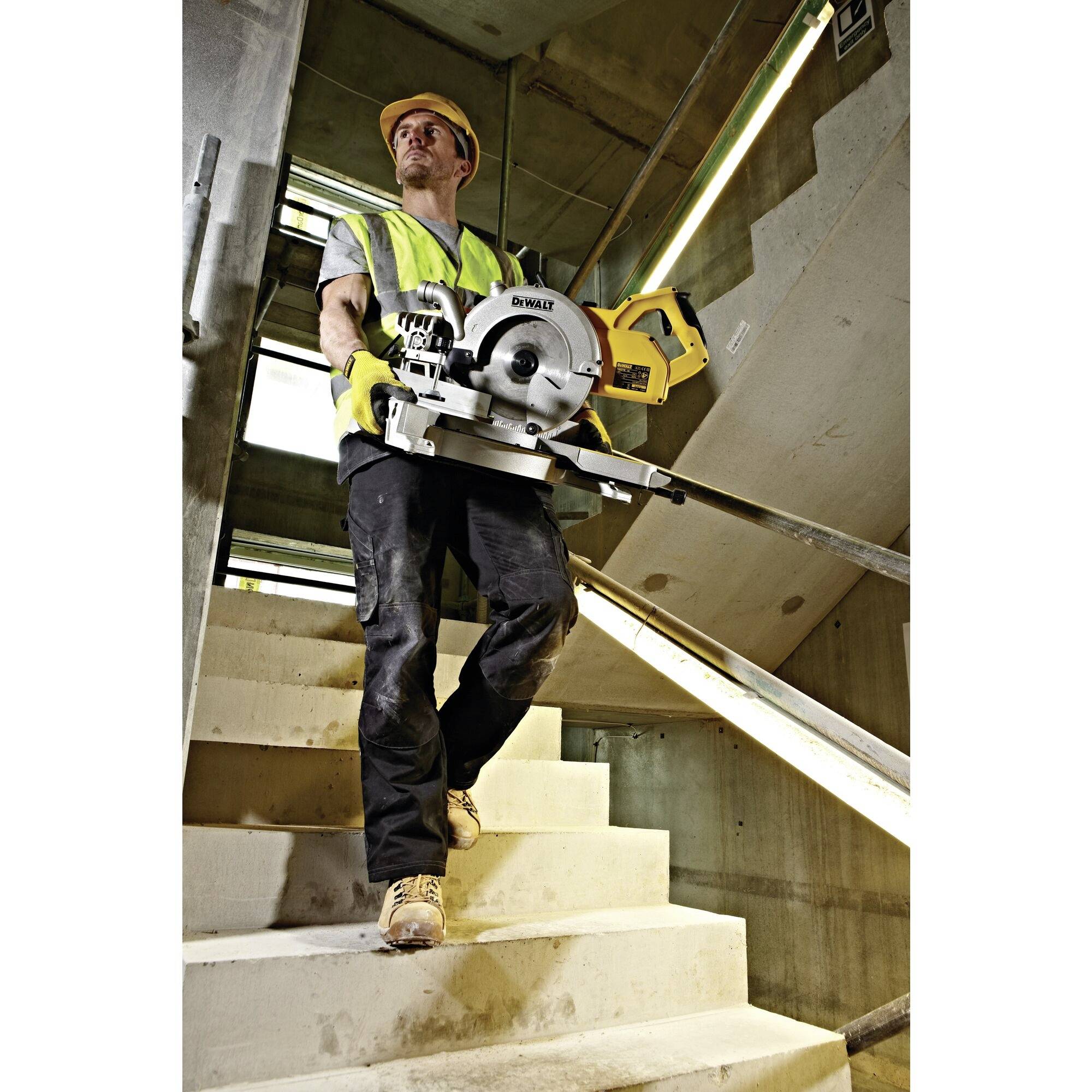 A worker in protective clothing carries a circular saw on a construction site staircase.