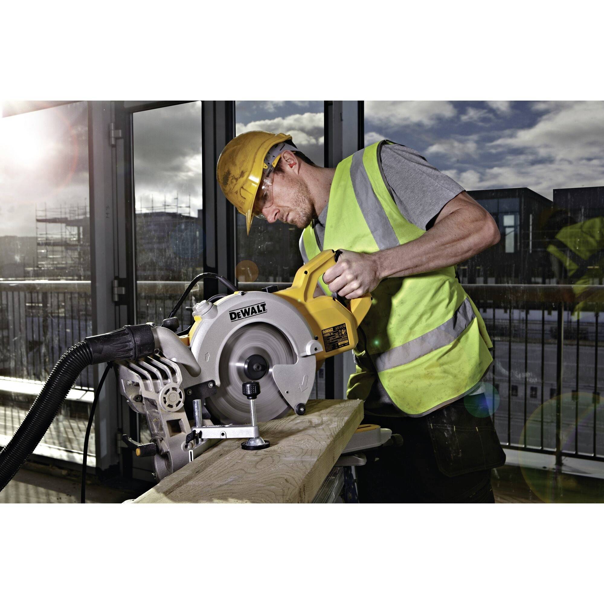 A construction worker wearing a hard hat and high-visibility vest is using an electric saw to cut a wooden plank.