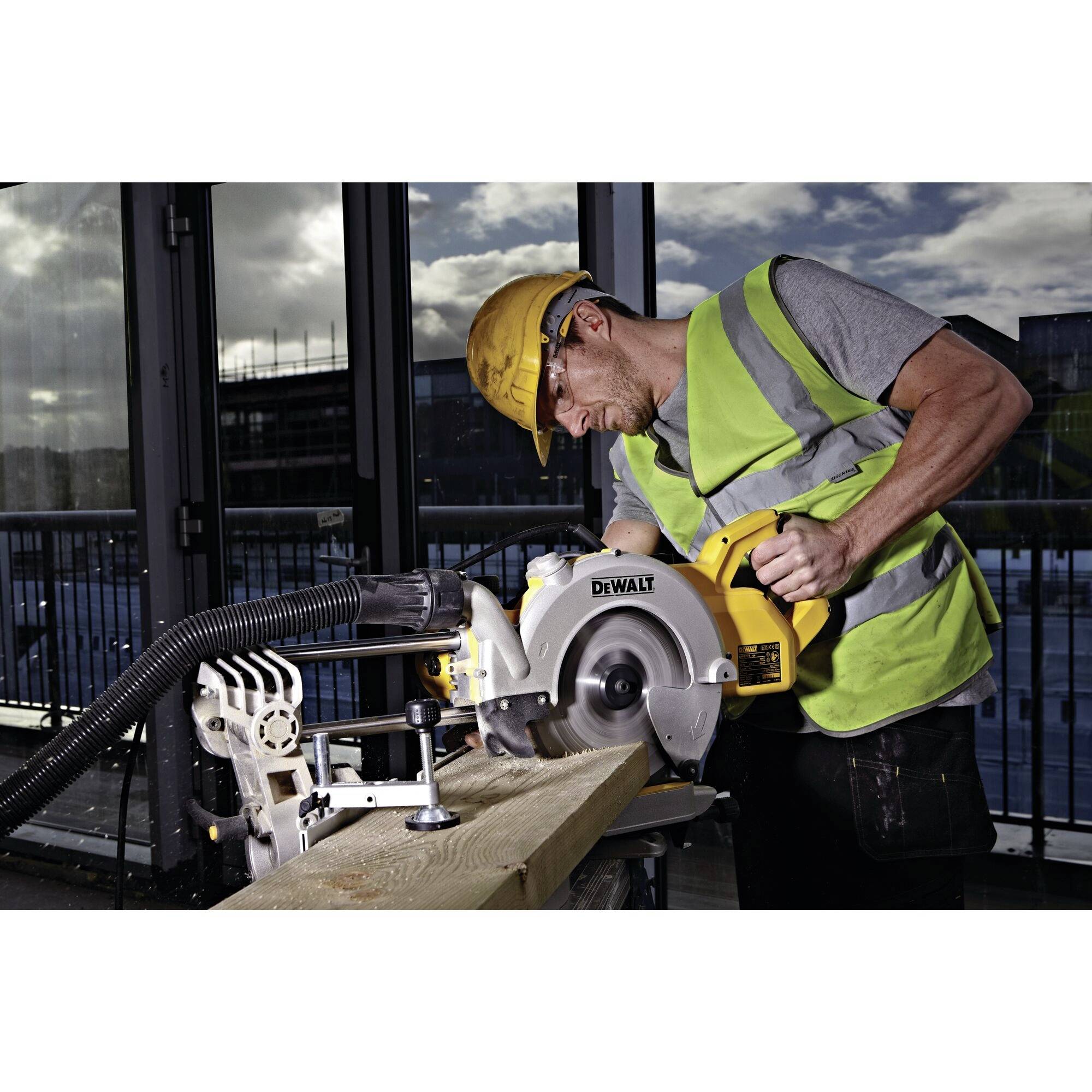 A person wearing a hard hat and high-visibility vest is cutting a wooden board with an electric circular saw in an industrial building.