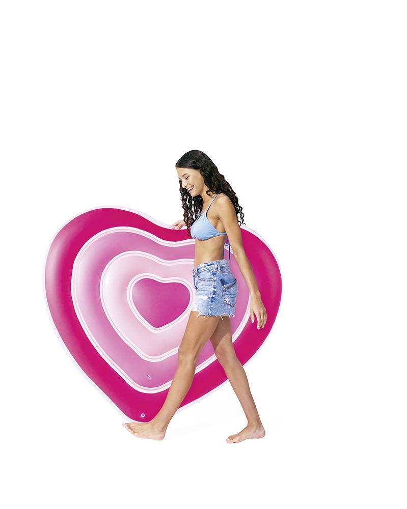 A woman in a blue bikini top and denim shorts is carrying a large, heart-shaped, pink and white swimming float against a white background.