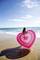 A woman on the beach holds a heart-shaped inflatable ring, while small waves caress the sand. The sky is clear and sunny.