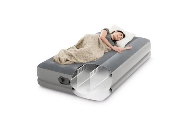 A person is sleeping on a grey air mattress, wrapped in a blanket. A cross-section reveals the internal structure of the mattress.