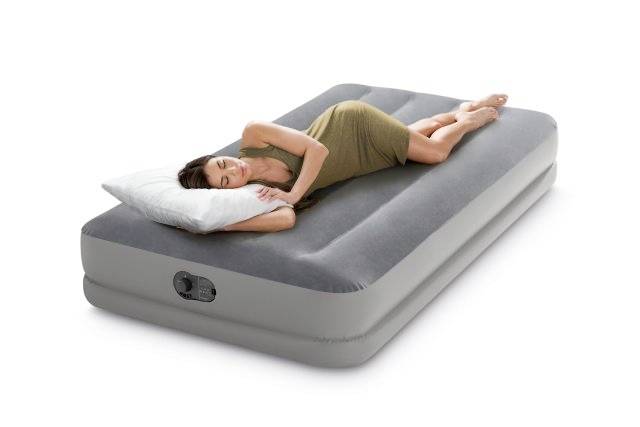 A woman is lying on an inflatable mattress, using a pillow, looking relaxed. The mattress is grey, rectangular, with an integrated valve.