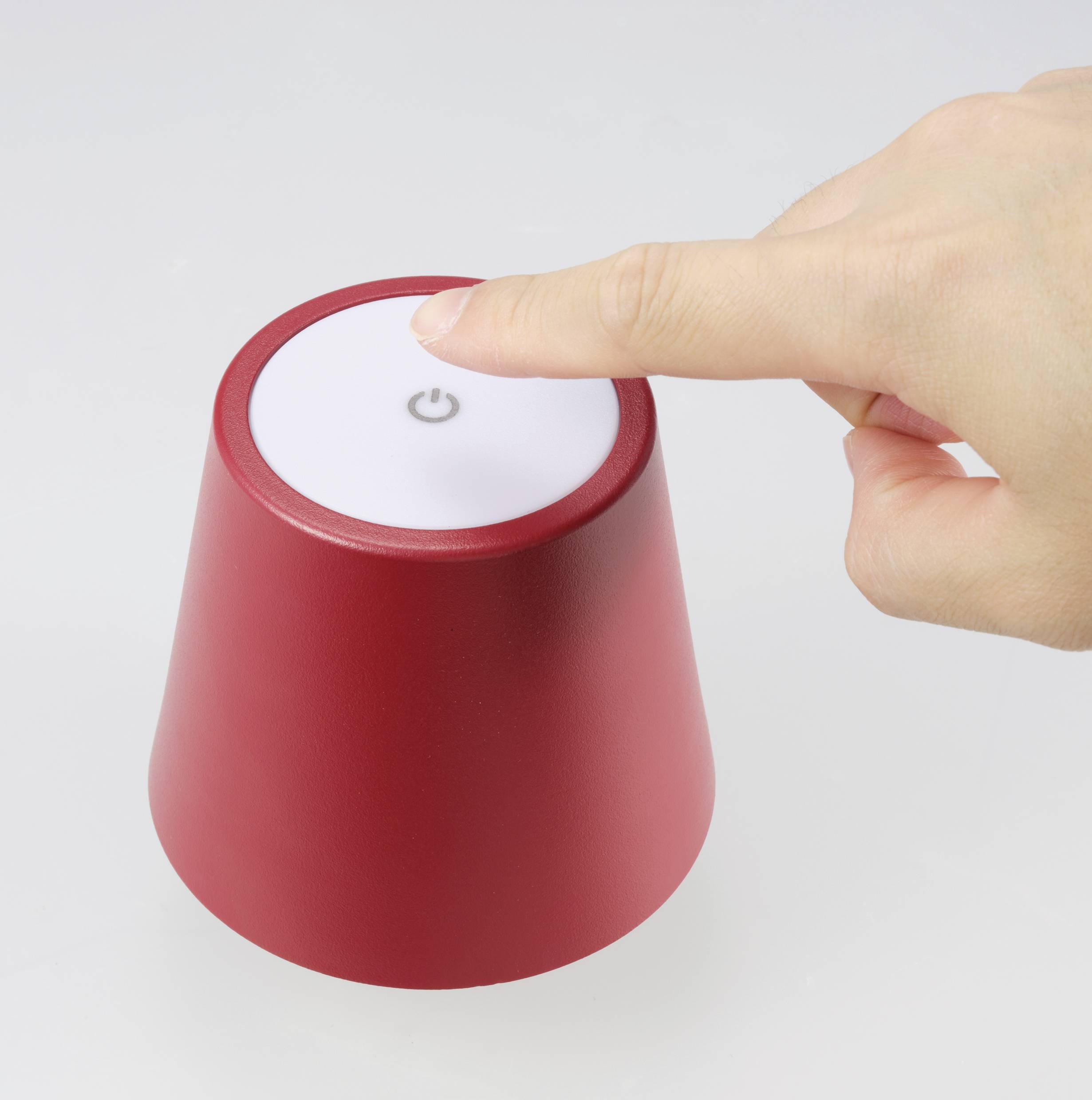 A hand presses the top button of a small, red lamp to switch it on or off. The lamp is situated on a white surface.