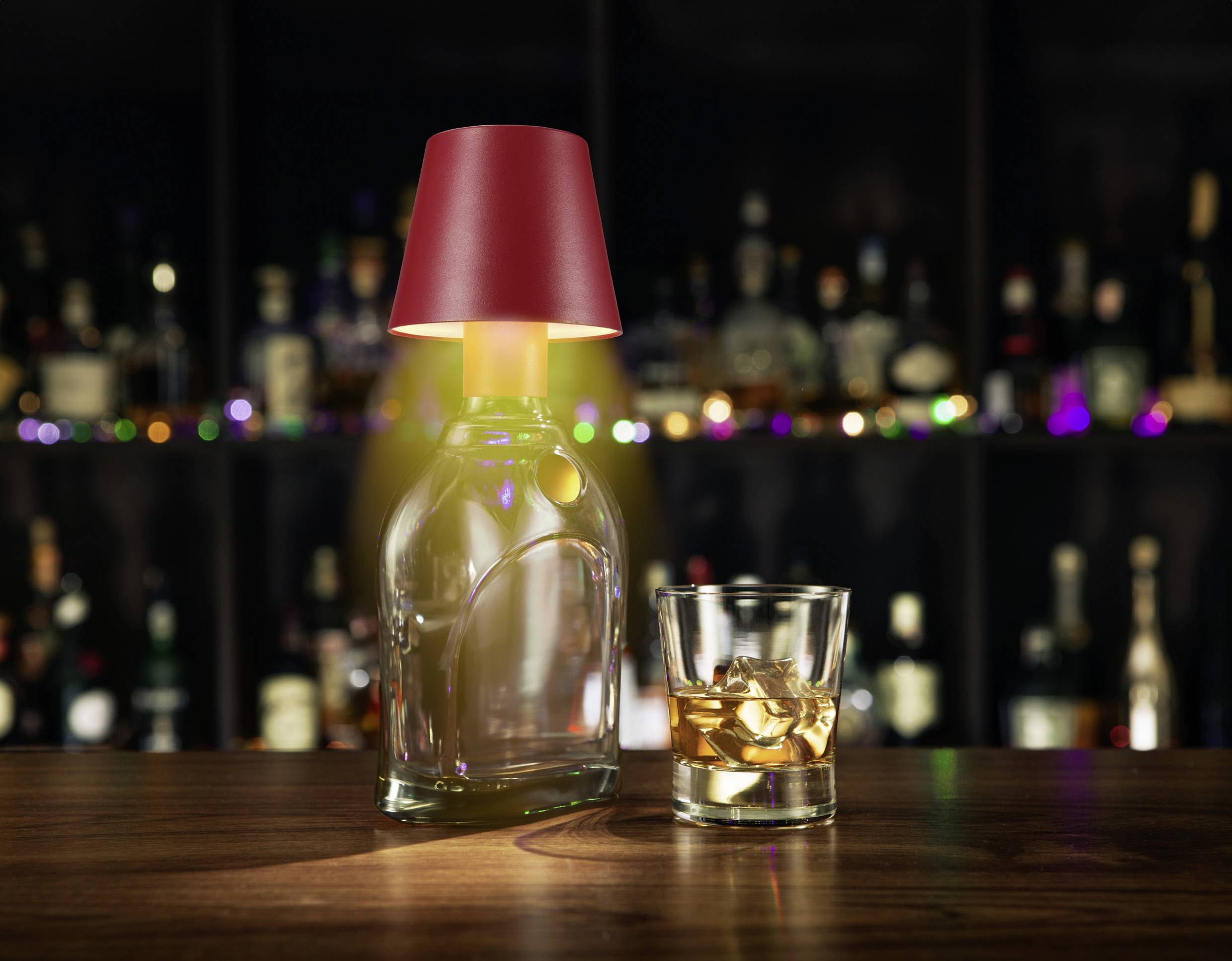 A glass bottle with a red lampshade and a glass of whisky with ice cubes sit on a wooden bar counter in front of a bar.