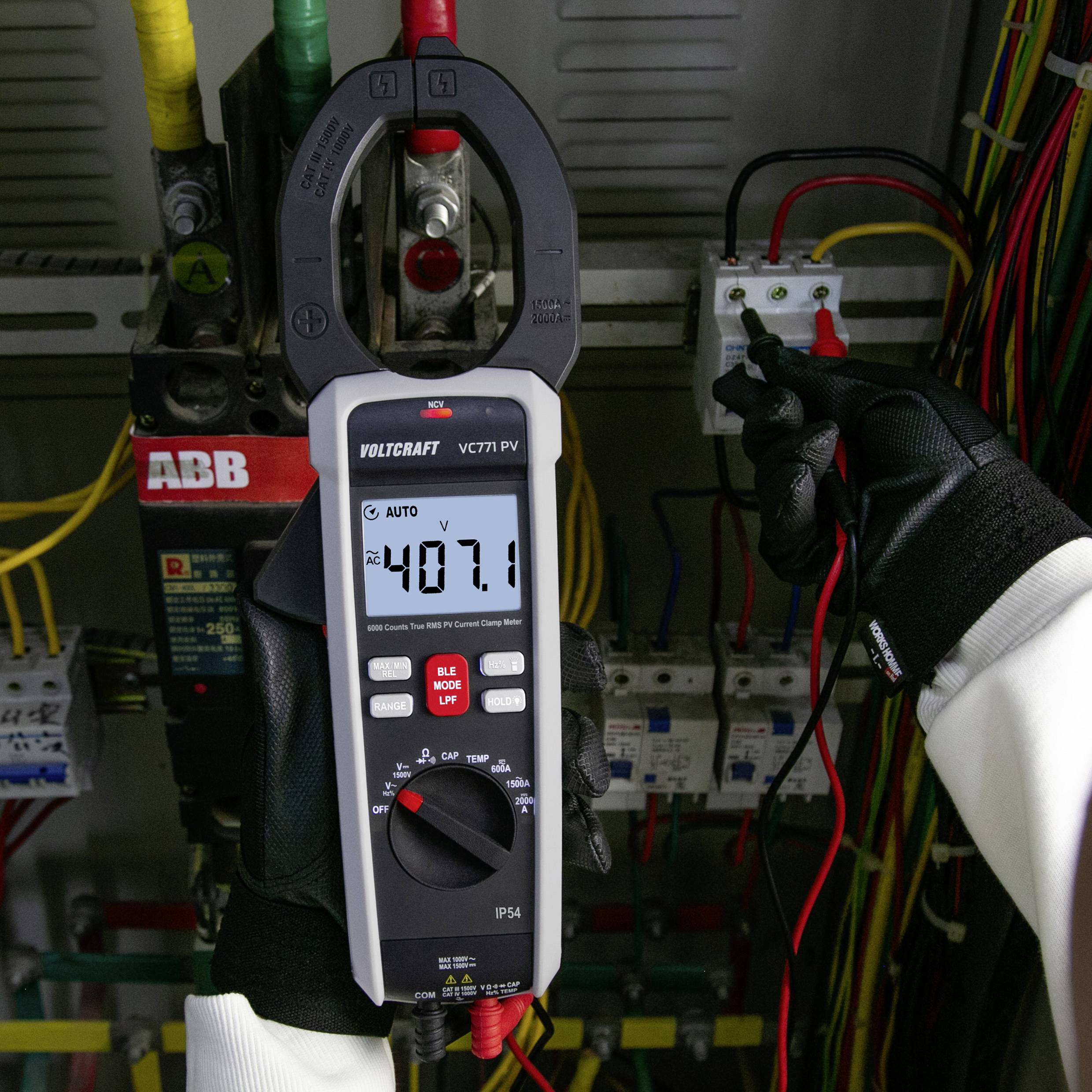 A person wearing gloves uses a multimeter to measure voltage, showing '407.1' on the display, in front of a control panel with wires.