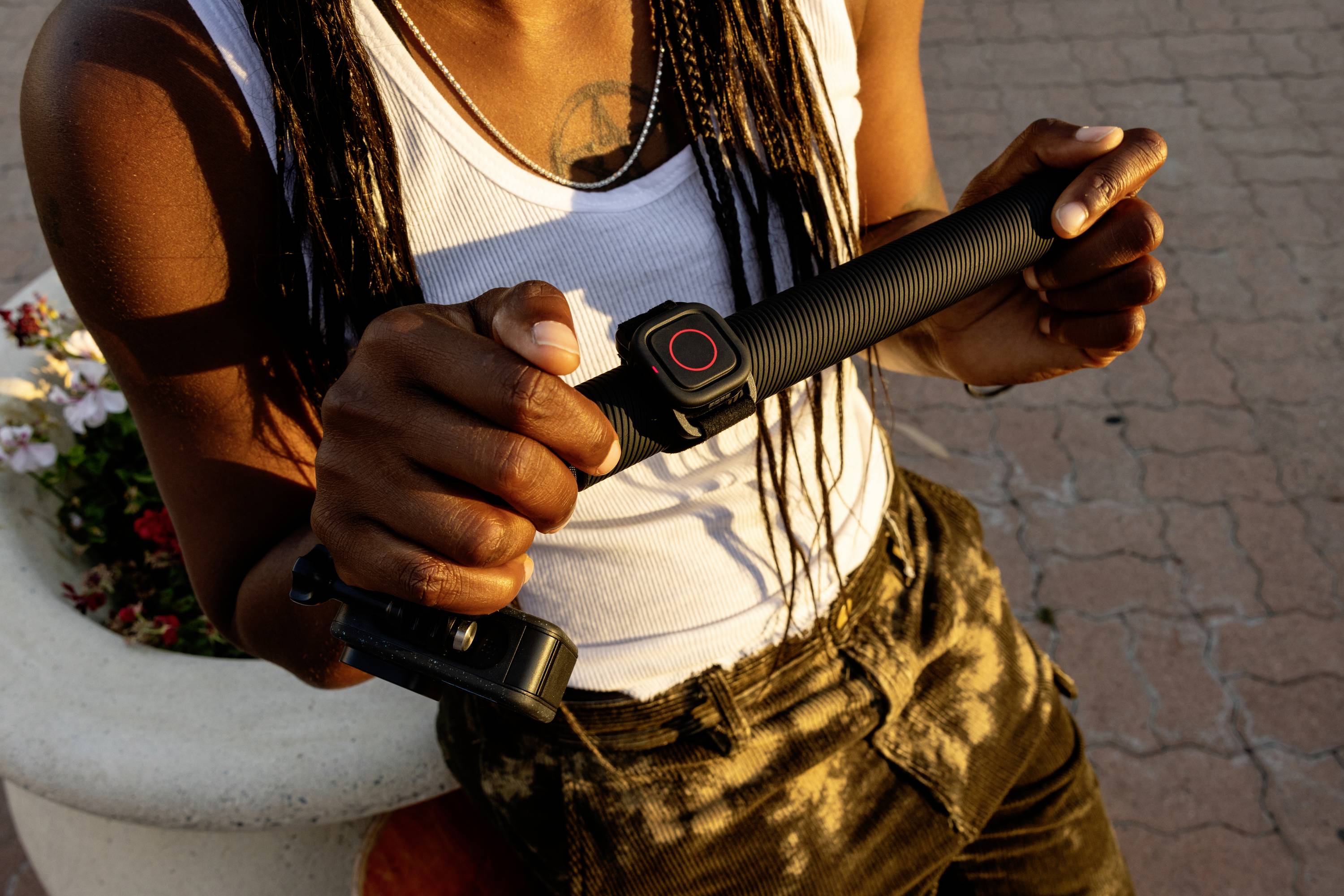 A person is holding a monopod grip with a camera, standing beside a flower bed on a paved surface.