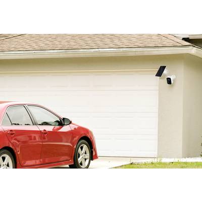 Red car parked in front of a closed, white garage. A surveillance camera with a solar panel is mounted on the exterior wall.