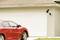 Red car parked in front of a closed, white garage. A surveillance camera with a solar panel is mounted on the exterior wall.