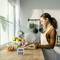 A woman is chopping onions in a modern kitchen. On the table are a bowl of tomatoes, a tray of citrus fruits, and a kitchen timer.