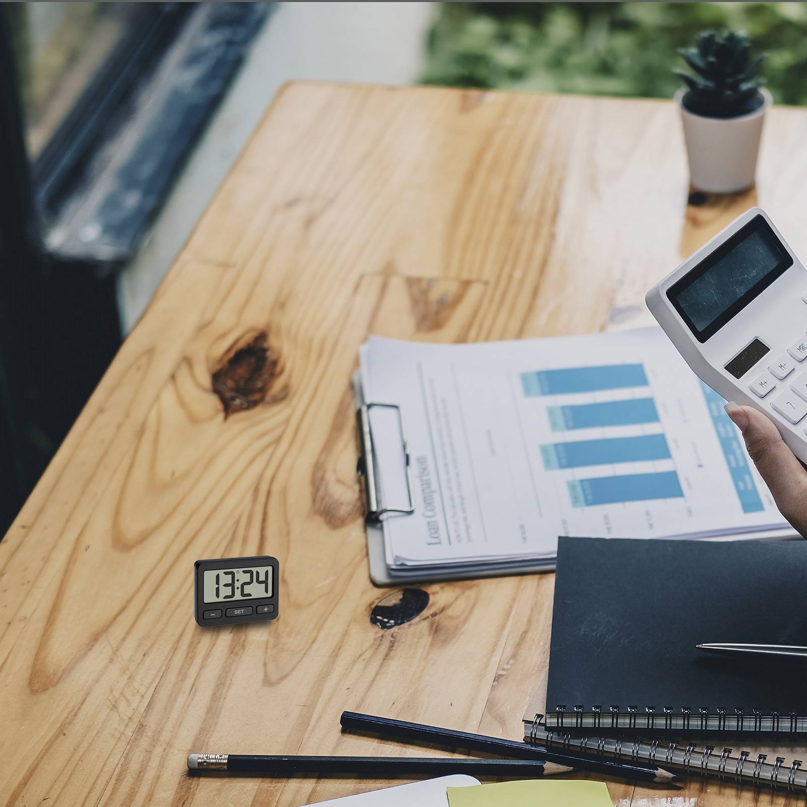 A calculator, notebook and a diagram with blue bars are lying on a table. A clock shows 13:24. A plant is in the background.