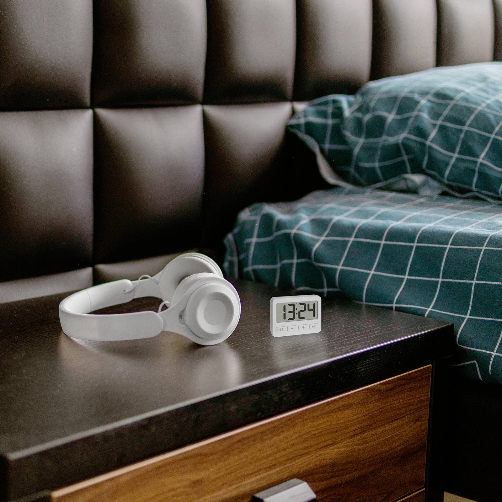 Alarm clock shows 13:24 on a bedside table next to headphones and a bed with a blue-checked pillow.