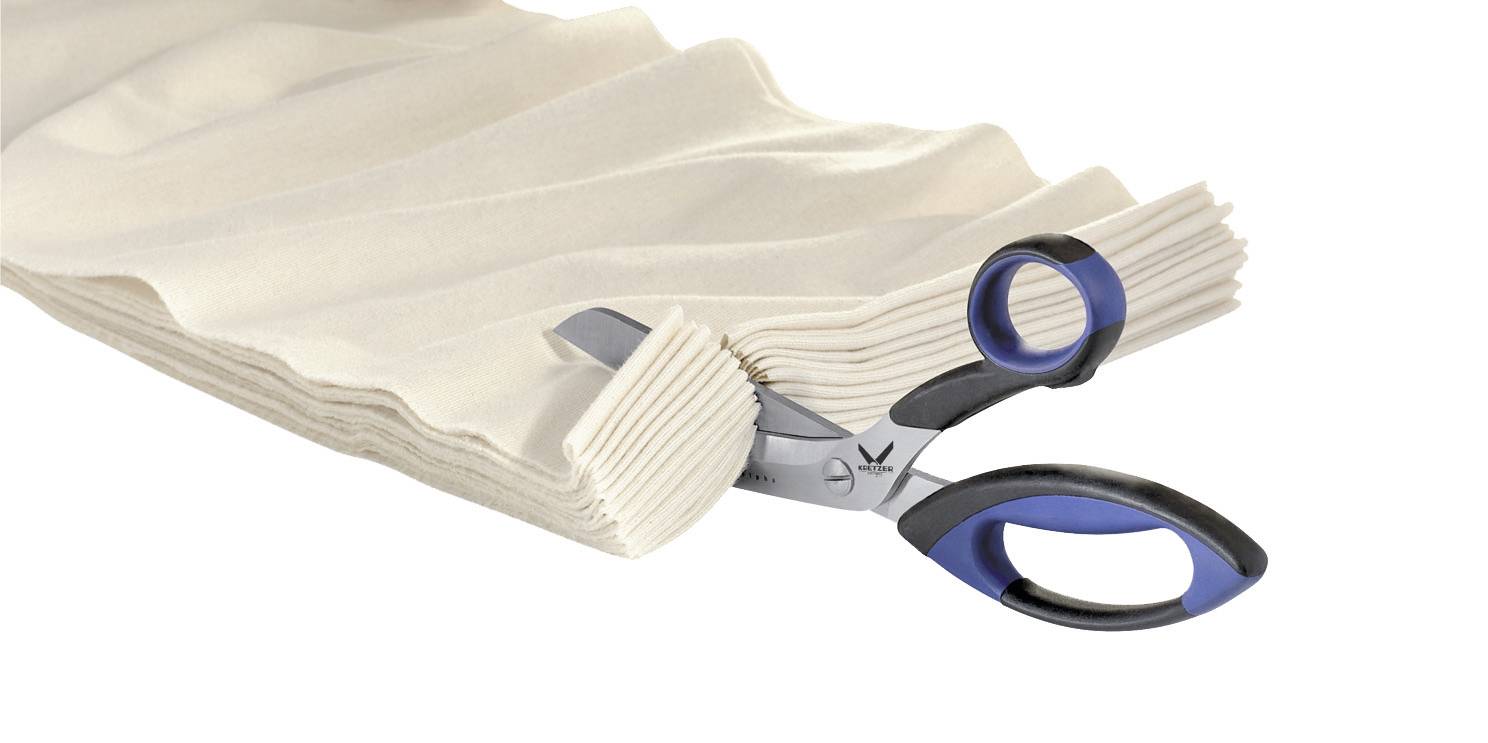 A pair of scissors cuts through several layers of beige fabric. The fabric is arranged in a wavy pattern, and the scissors have blue handles.