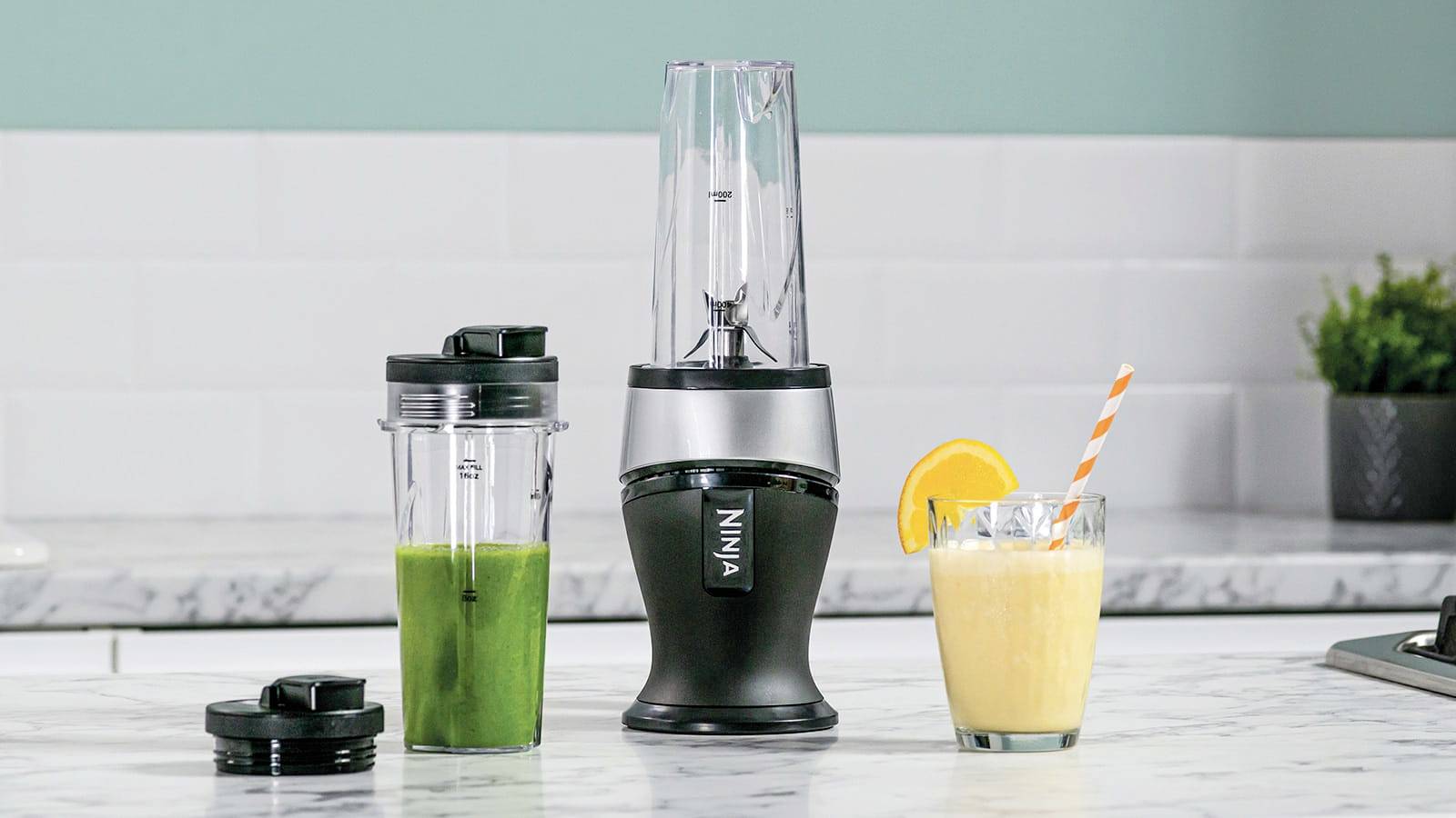 A blender on a kitchen worktop with a green smoothie in a cup and a yellow drink with an orange slice and a straw.