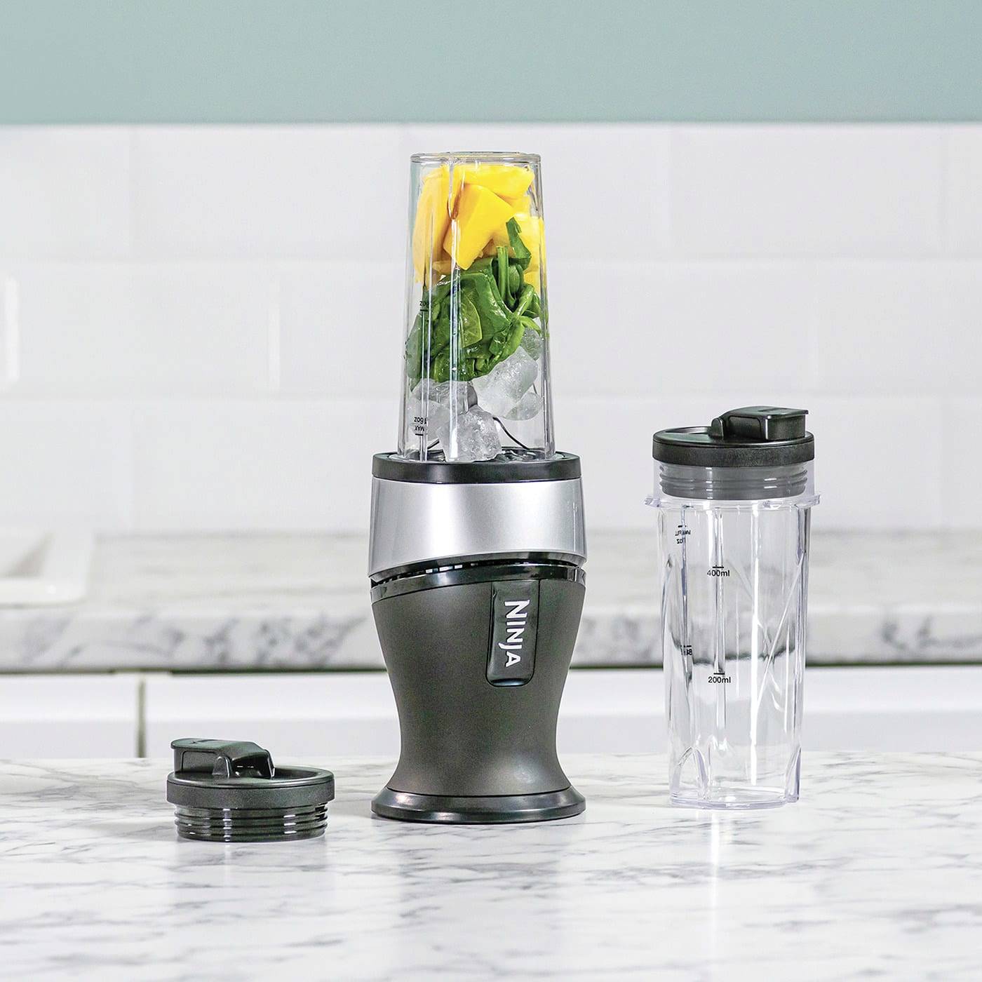 A blender filled with spinach, mango, and ice stands on a kitchen worktop, with an empty additional container beside it.