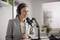 A smiling woman in a light grey blazer is speaking into a microphone. She is wearing headphones and sitting in a modern room.