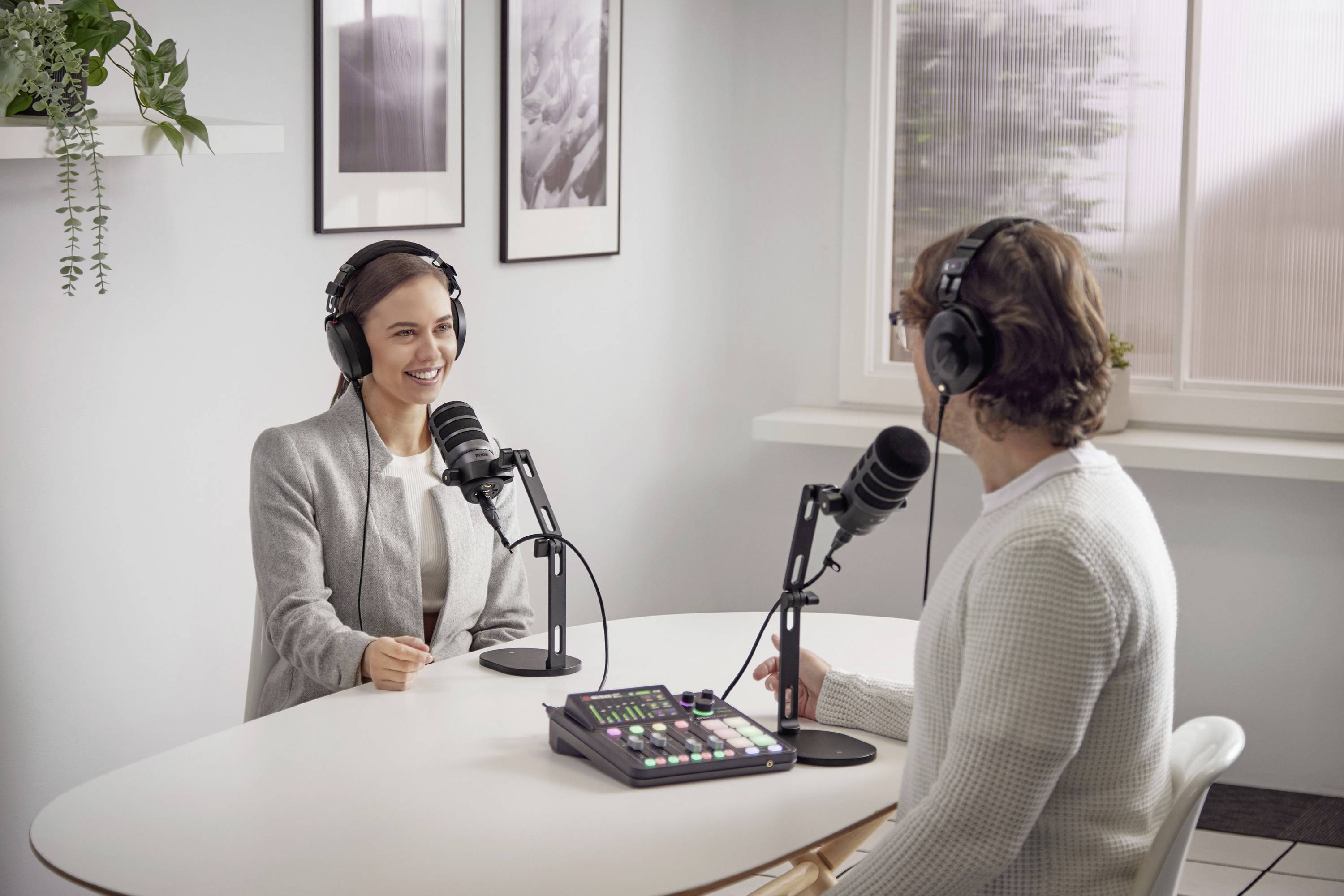 Two people with headphones are conversing at a table with microphones. Pictures are hanging on the wall in the background.