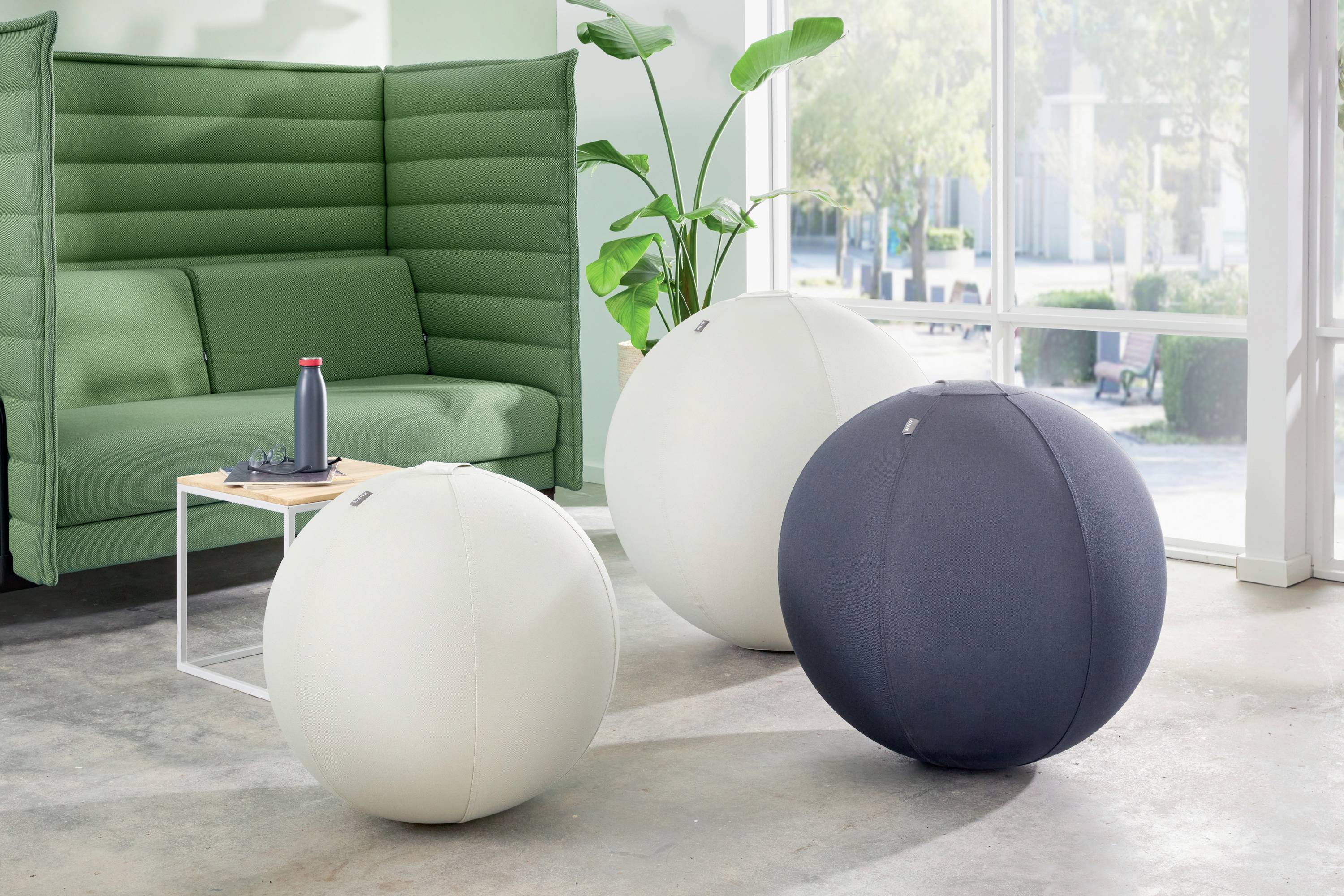 Three large, round exercise balls in the foreground of a modern, bright room, next to a green sofa with a small table and a plant.