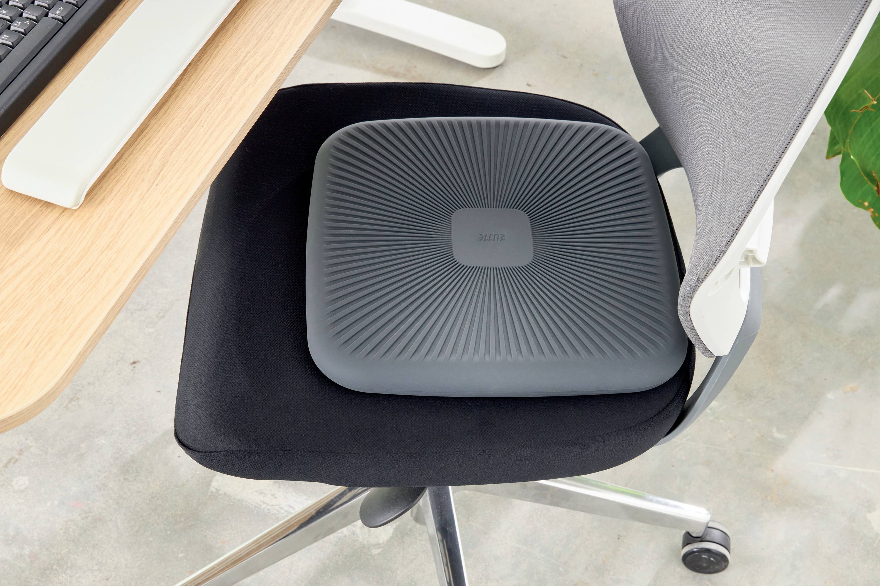 Seat cushion on an office chair next to a desk with a keyboard. The cushion is dark grey and square with a ribbed surface.