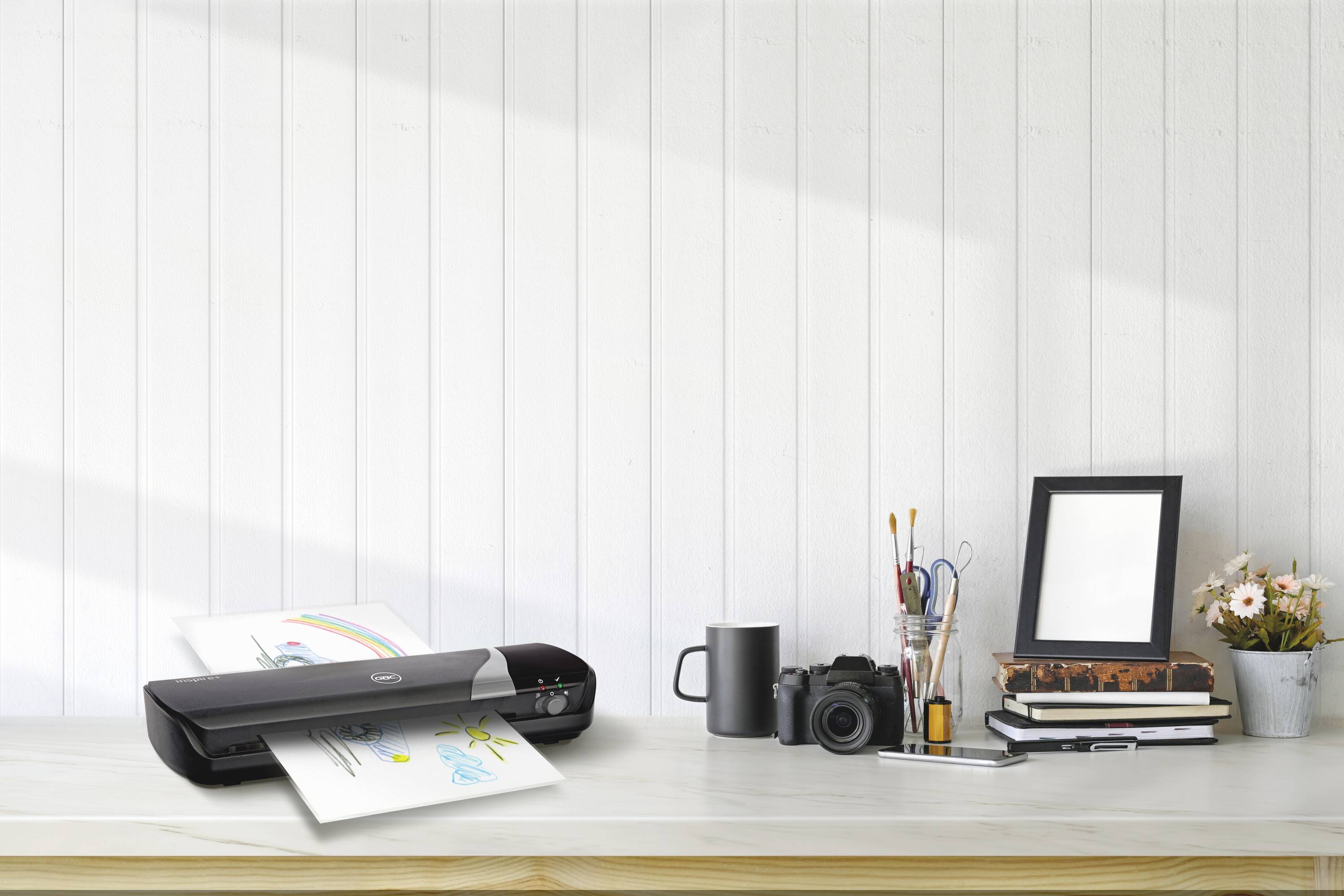 A printer on a desk is printing colourful documents. Beside the printer are a camera, books, flowers, and pens.