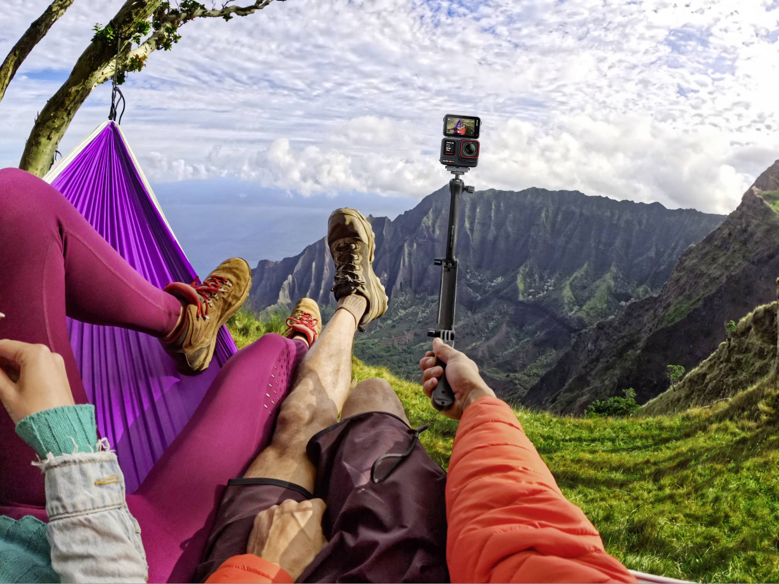 People are relaxing in a hammock with a mountain view. One person is holding a camera to capture the scenery.