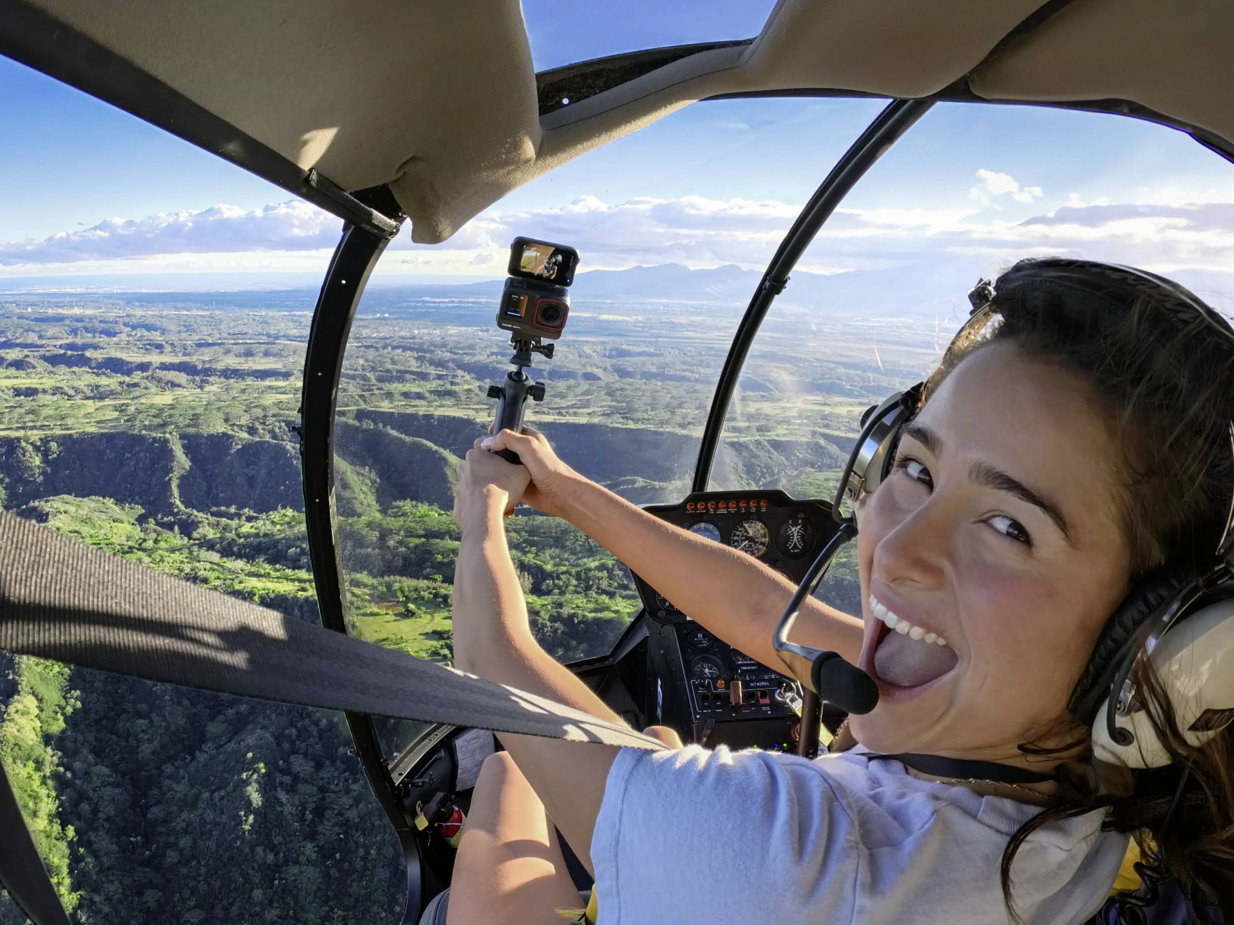 A smiling person is sitting in a helicopter, holding a GoPro camera and flying over a green landscape with mountains in the background.
