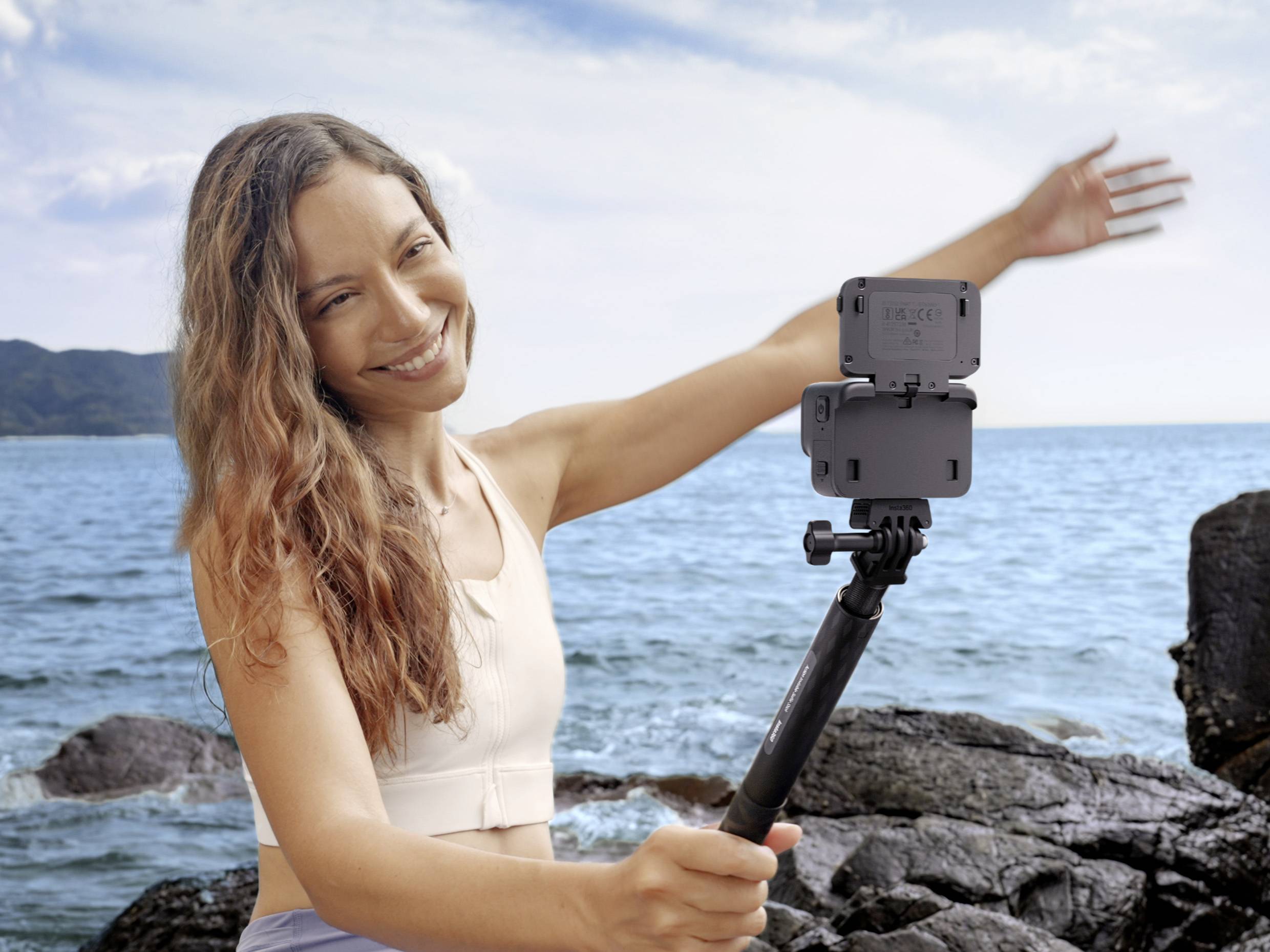 A woman is smiling while taking a selfie by the sea with a smartphone on a selfie stick. Rocks and water are visible in the background.