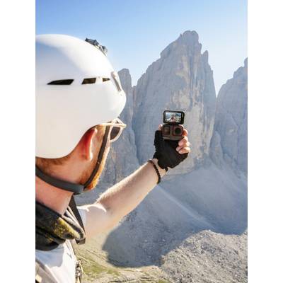 A person wearing a helmet takes a selfie with a camera, with the impressive rocks of the Dolomites against a clear sky in the background.