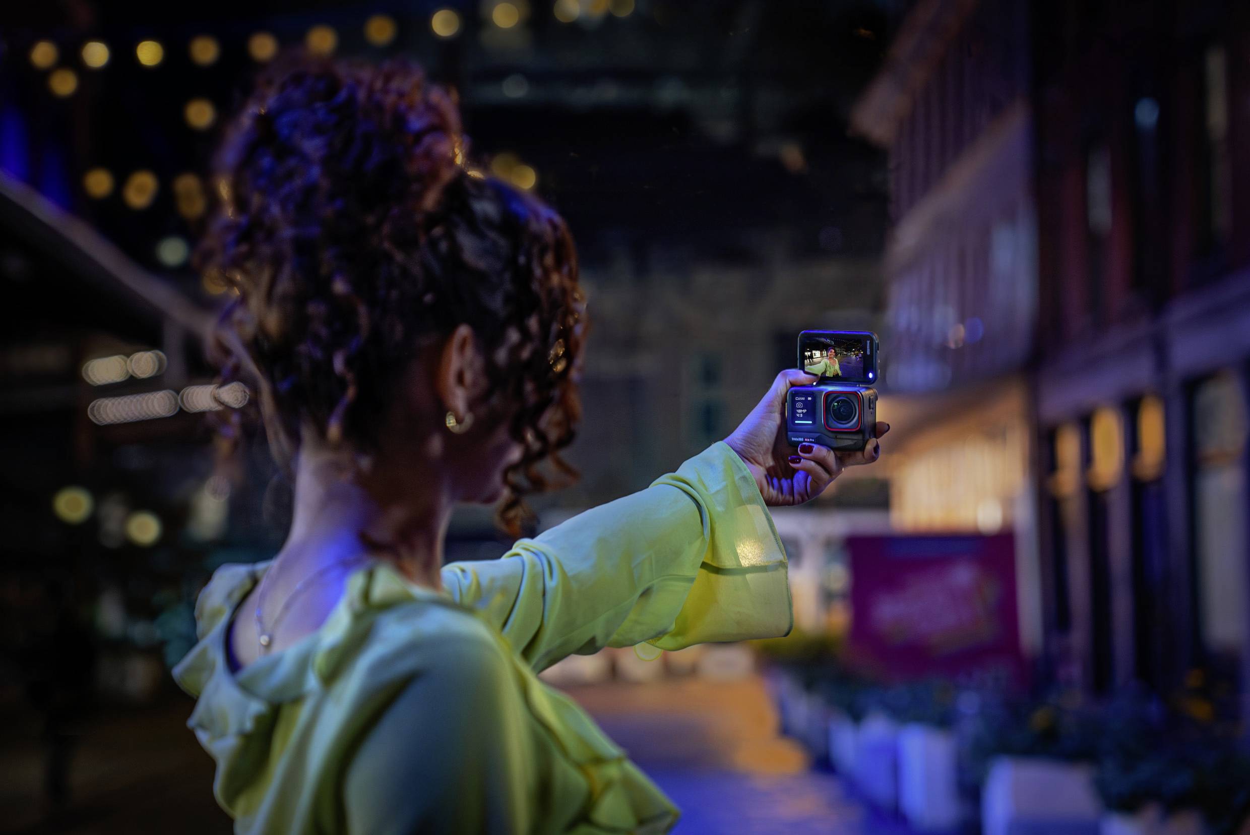 A woman with curly hair photographs herself at night in a lit-up city, camera in hand.