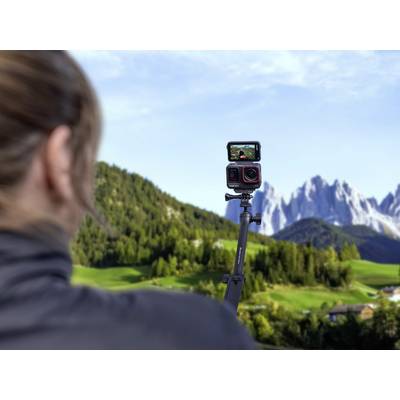 A person is filming with a camera on a tripod, pointed towards a mountainous landscape. Forests and mountains are visible in the background.