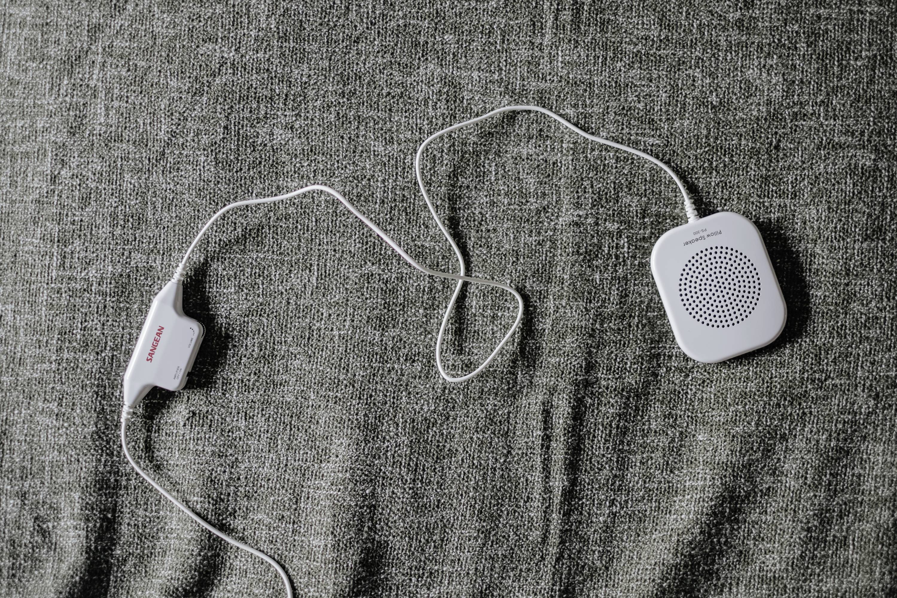 White portable speaker with long cables on a grey fabric background. Branded 'Sangean'.