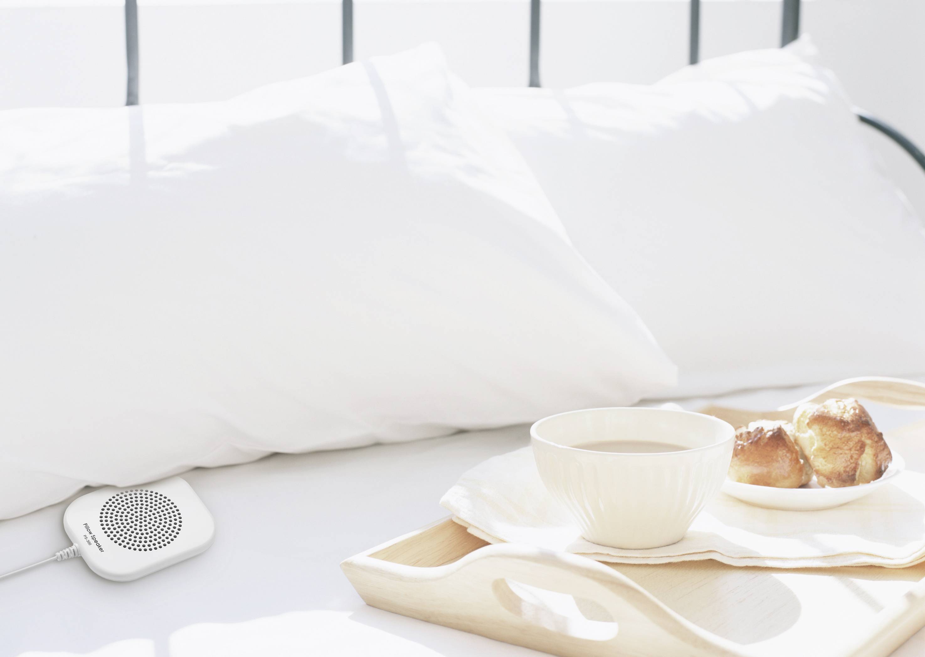 A tray with a cup of coffee and a croissant is lying on a white bed. A small speaker stands next to the bed.