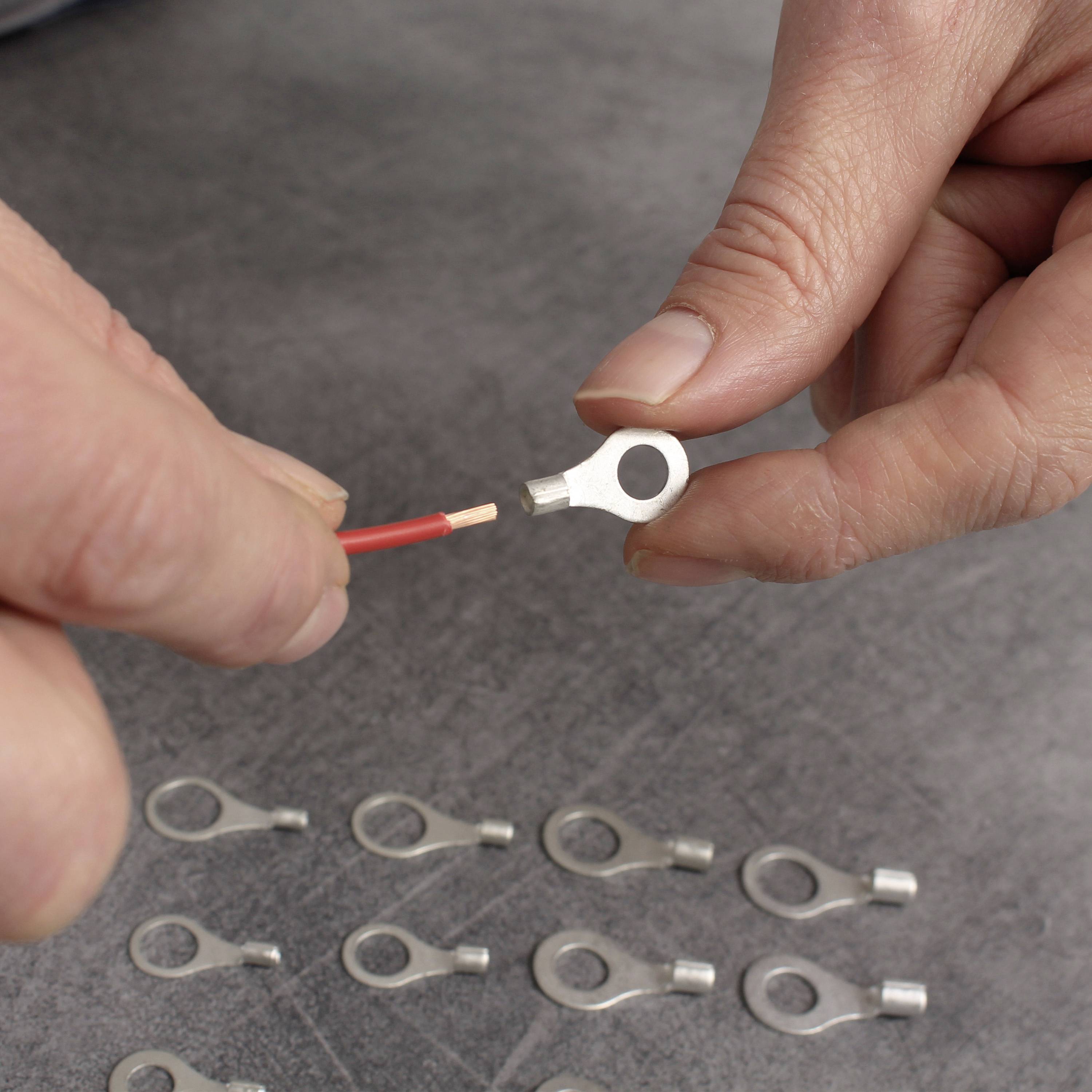 Two hands demonstrate how a cable end is being inserted into a metal ring cable lug. Several additional cable lugs are lying on the table.
