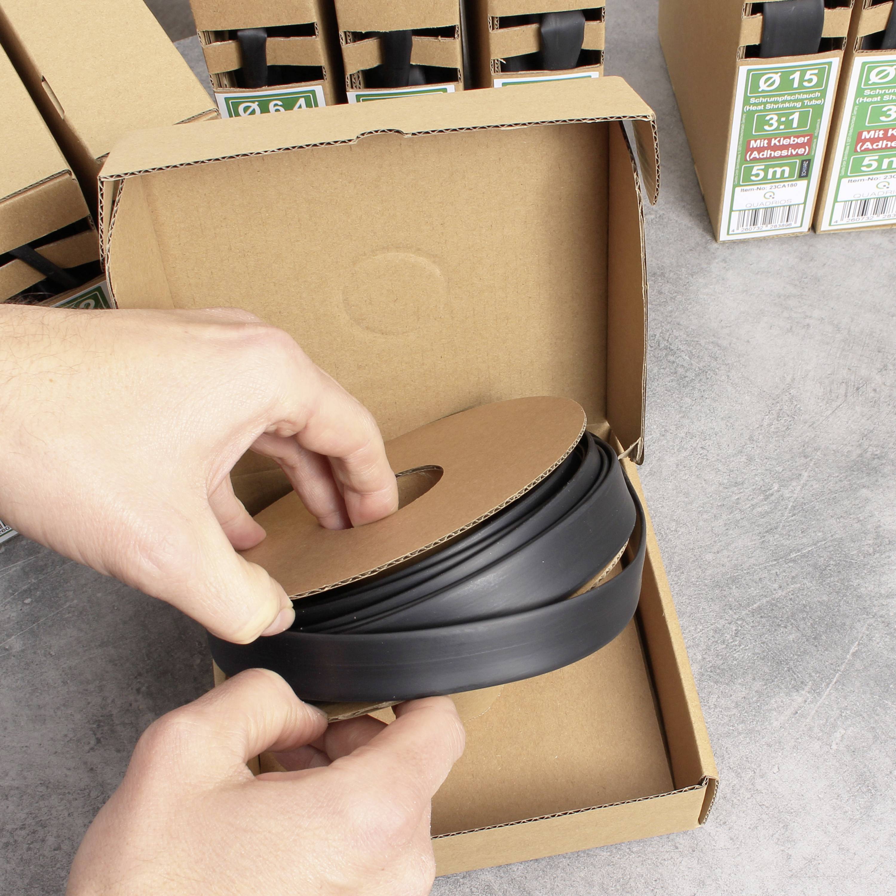A person is holding a roll of black cable ties in their hand, ready to place it into a cardboard box. In the background, additional boxes are stacked.