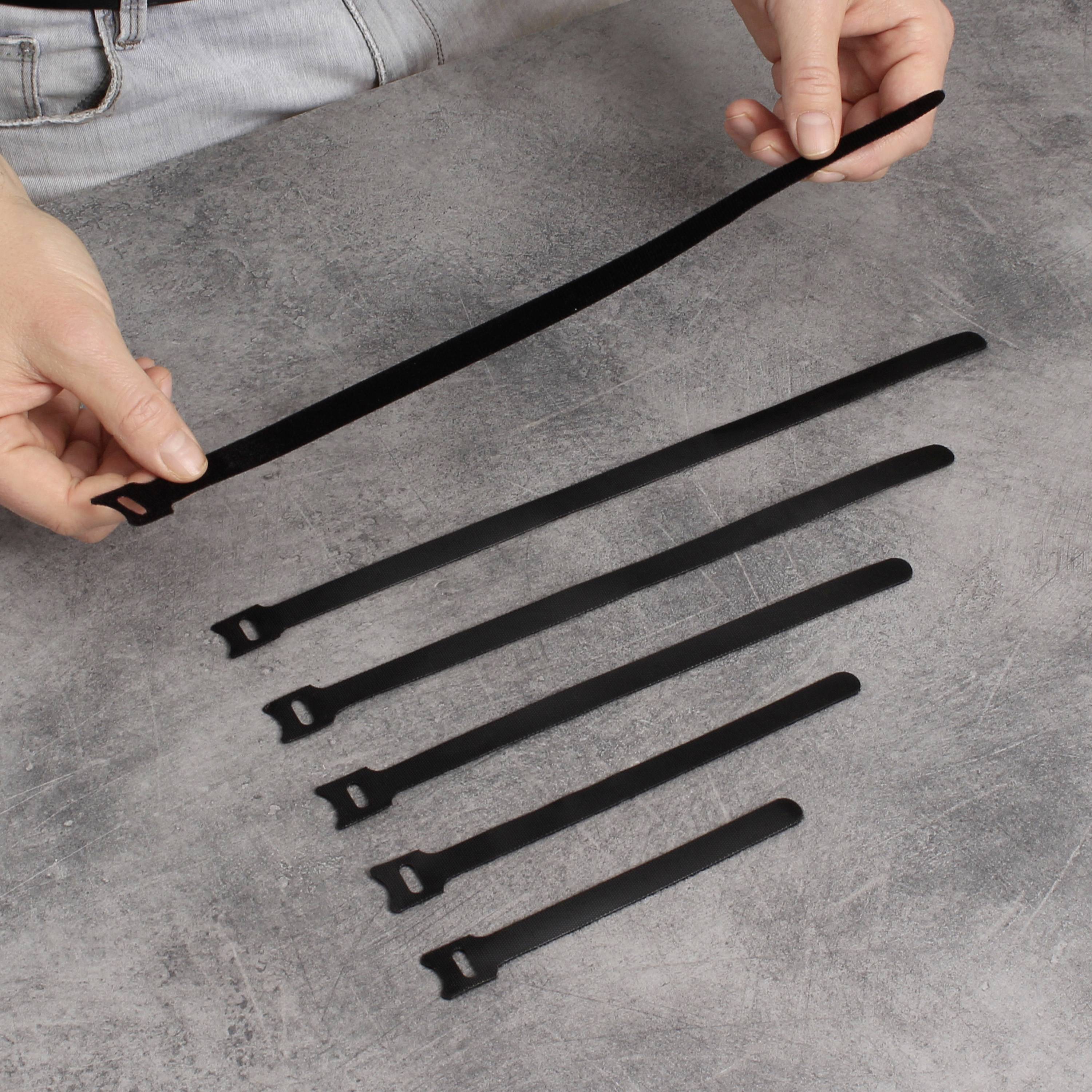 A person is holding a black cable tie above a grey surface, on which five additional cable ties of decreasing size are lying.