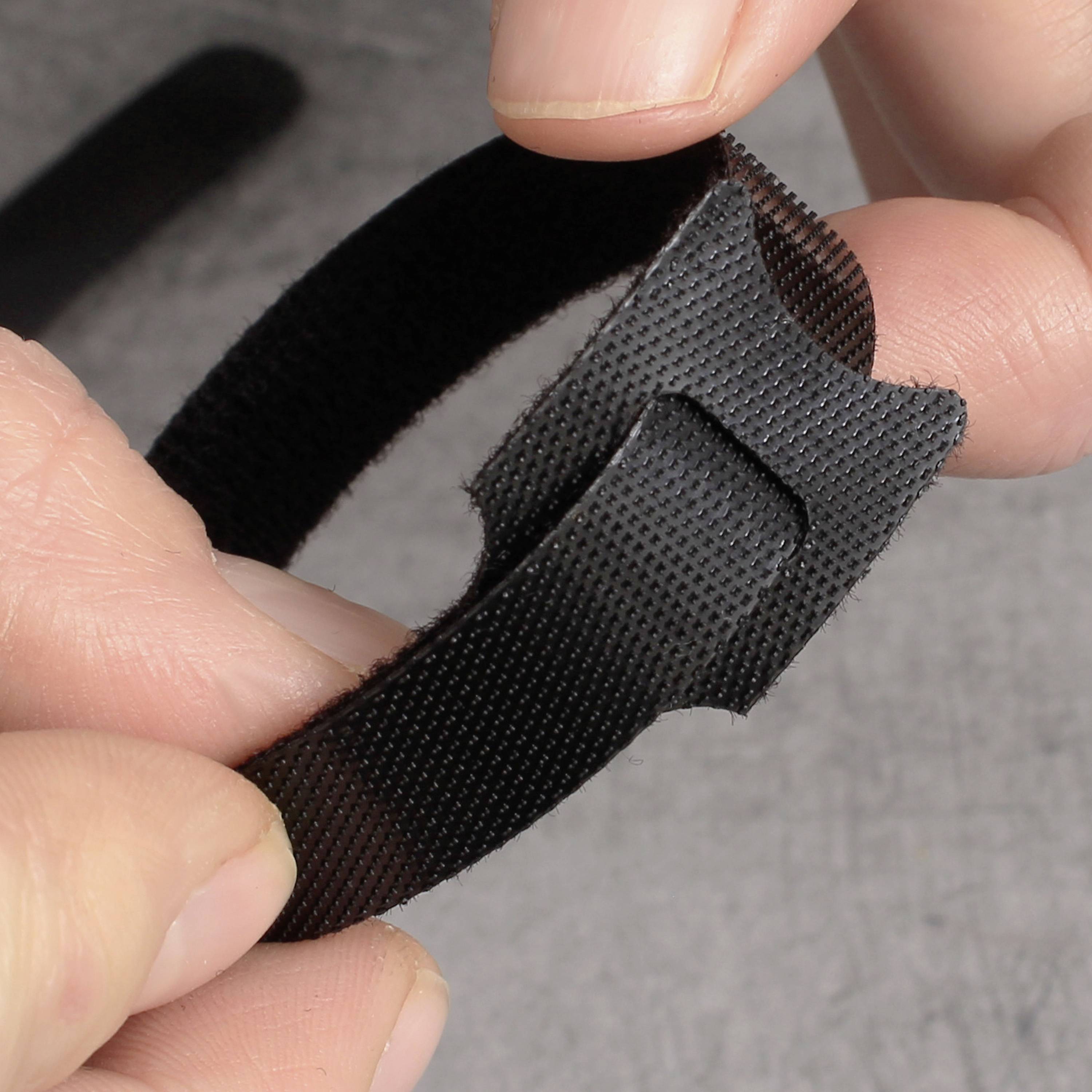 A hand is holding a black Velcro fastener that is slightly curved. The background is grey and out of focus.