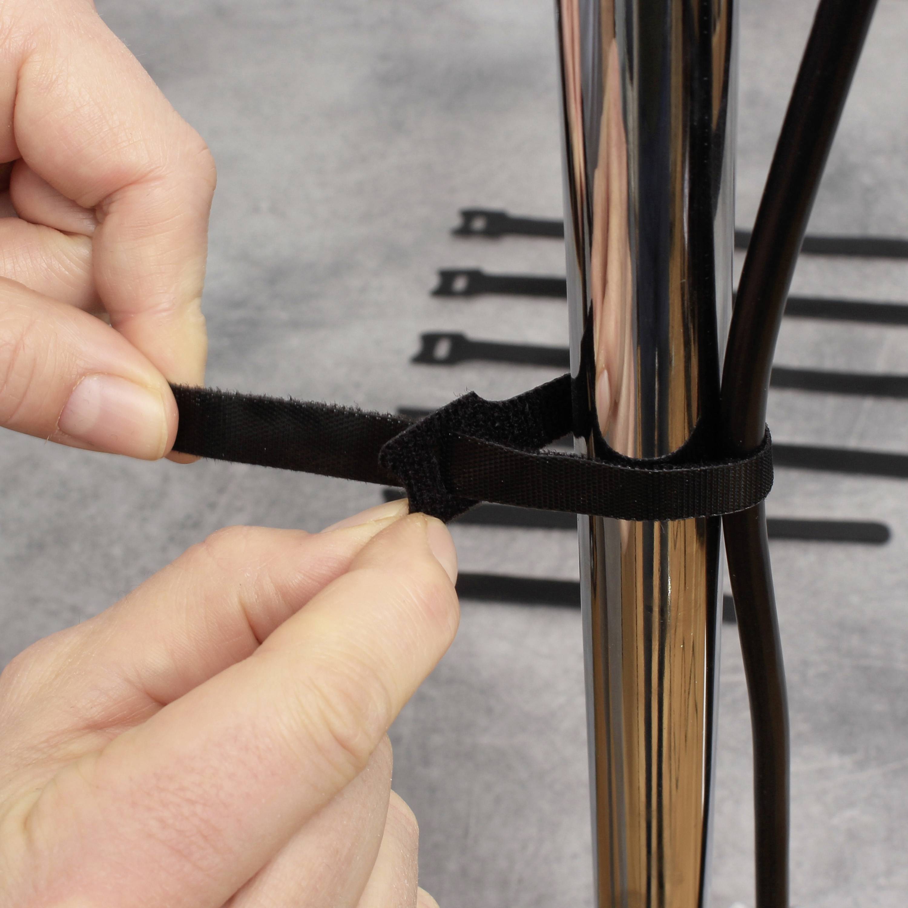 A hand is securing a cable with a hook-and-loop fastener around a metal pipe. Further hook-and-loop straps are visible in the background.