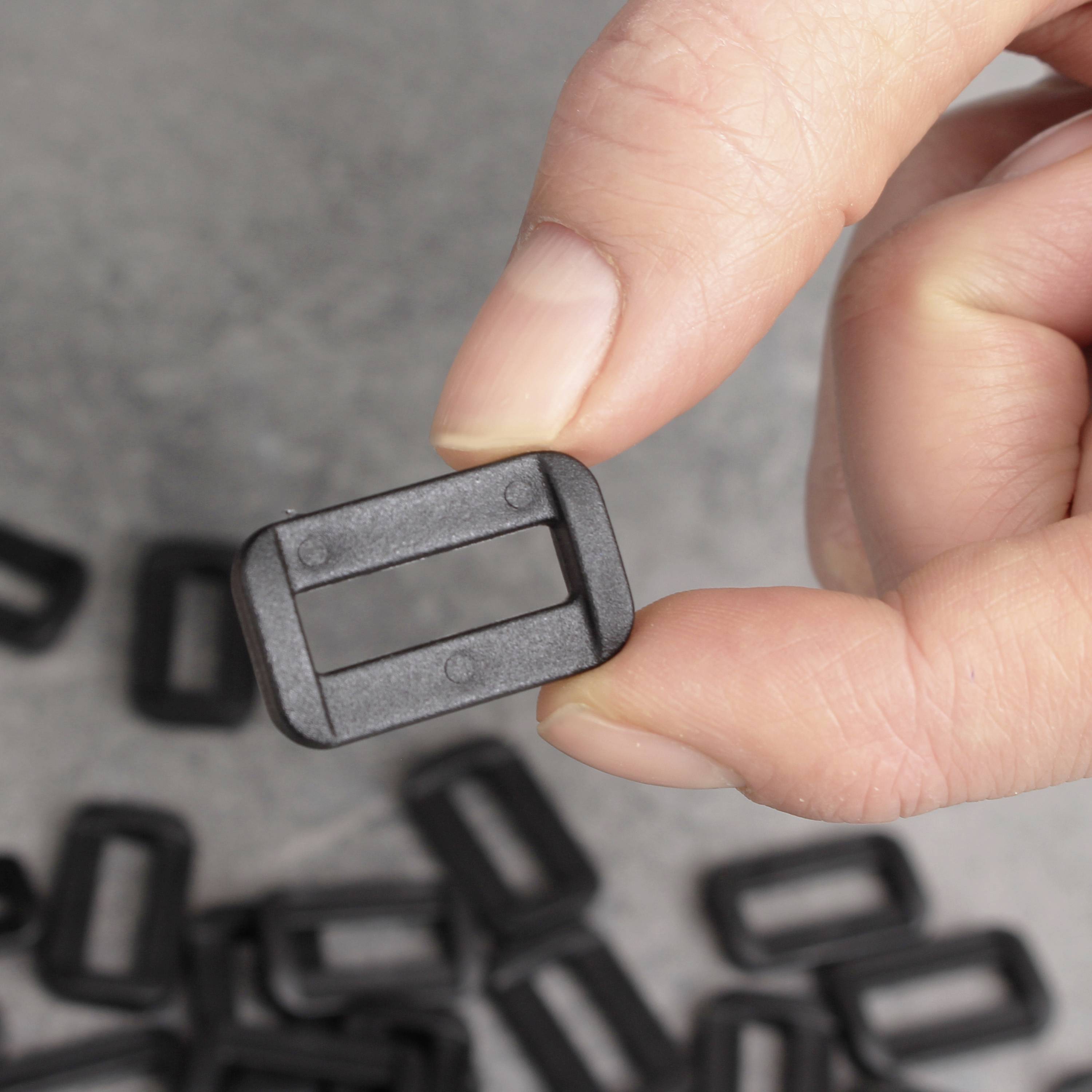 A hand is holding a small black rectangular plastic loop. In the background, further identical loops are distributed across a grey surface.