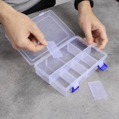 A person is inserting a divider into a transparent, empty plastic organiser with multiple compartments on a grey table.