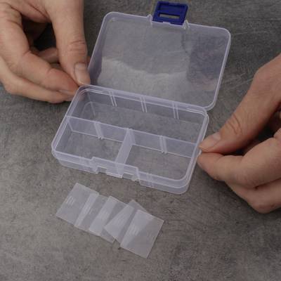 Two hands open a small transparent plastic container with multiple compartments. Four dividers lie beside it on a table.