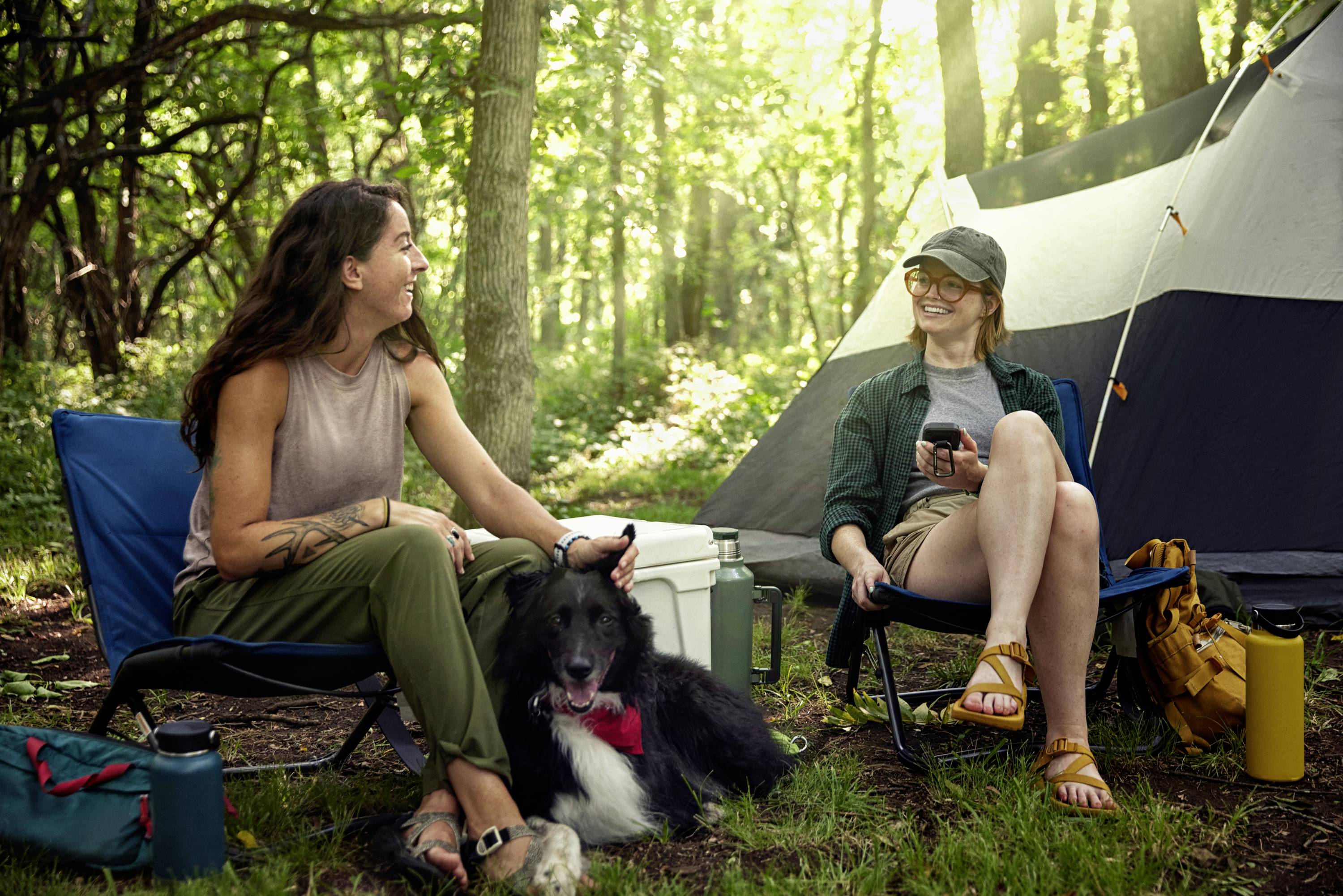 Two people are sitting relaxed on camping chairs in the woods next to a tent, they are laughing and have a black dog with them.