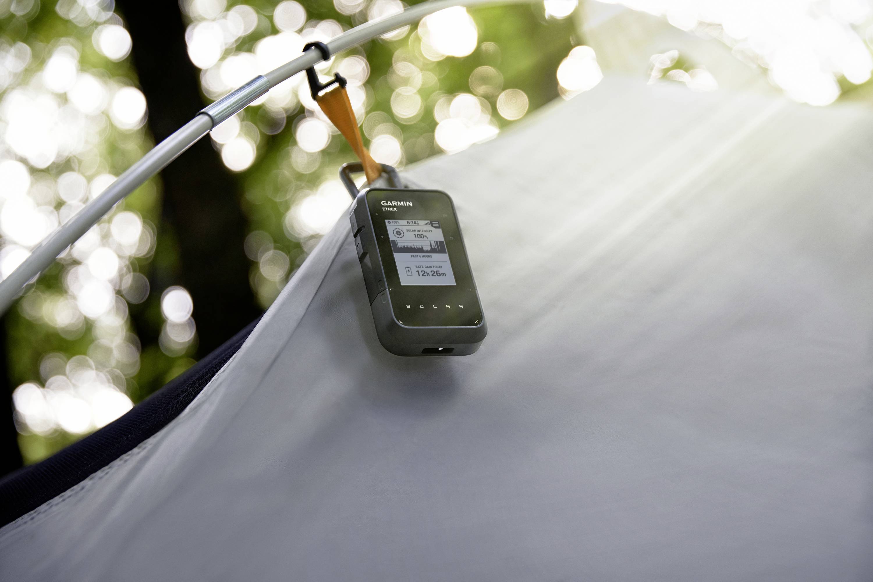 A GPS device is hanging on a tent pitched in a wooded area. Sunlight filters through the trees in the background.