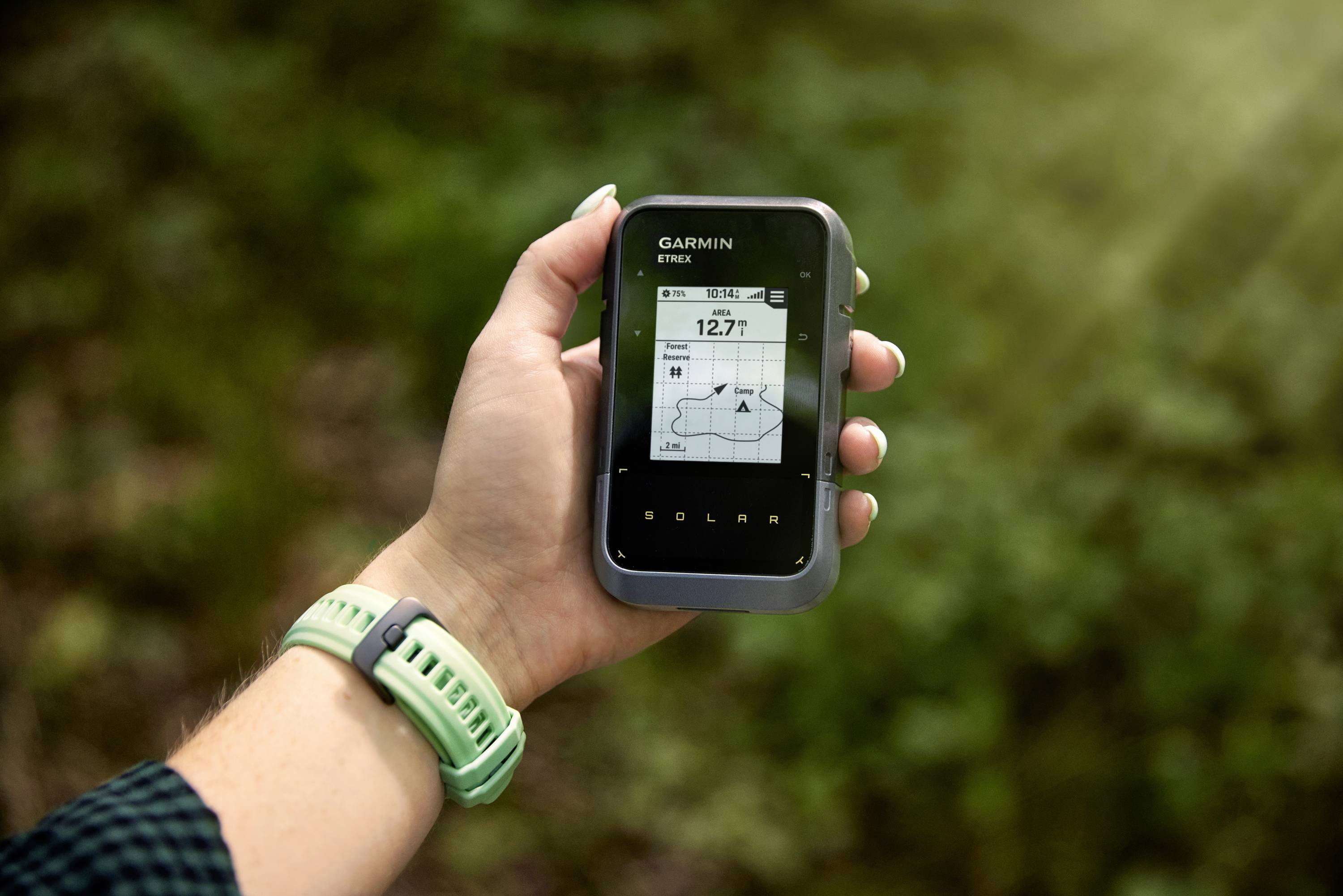 A hand is holding a GPS device in the woods, displaying a map with a distance of 12.7 km.