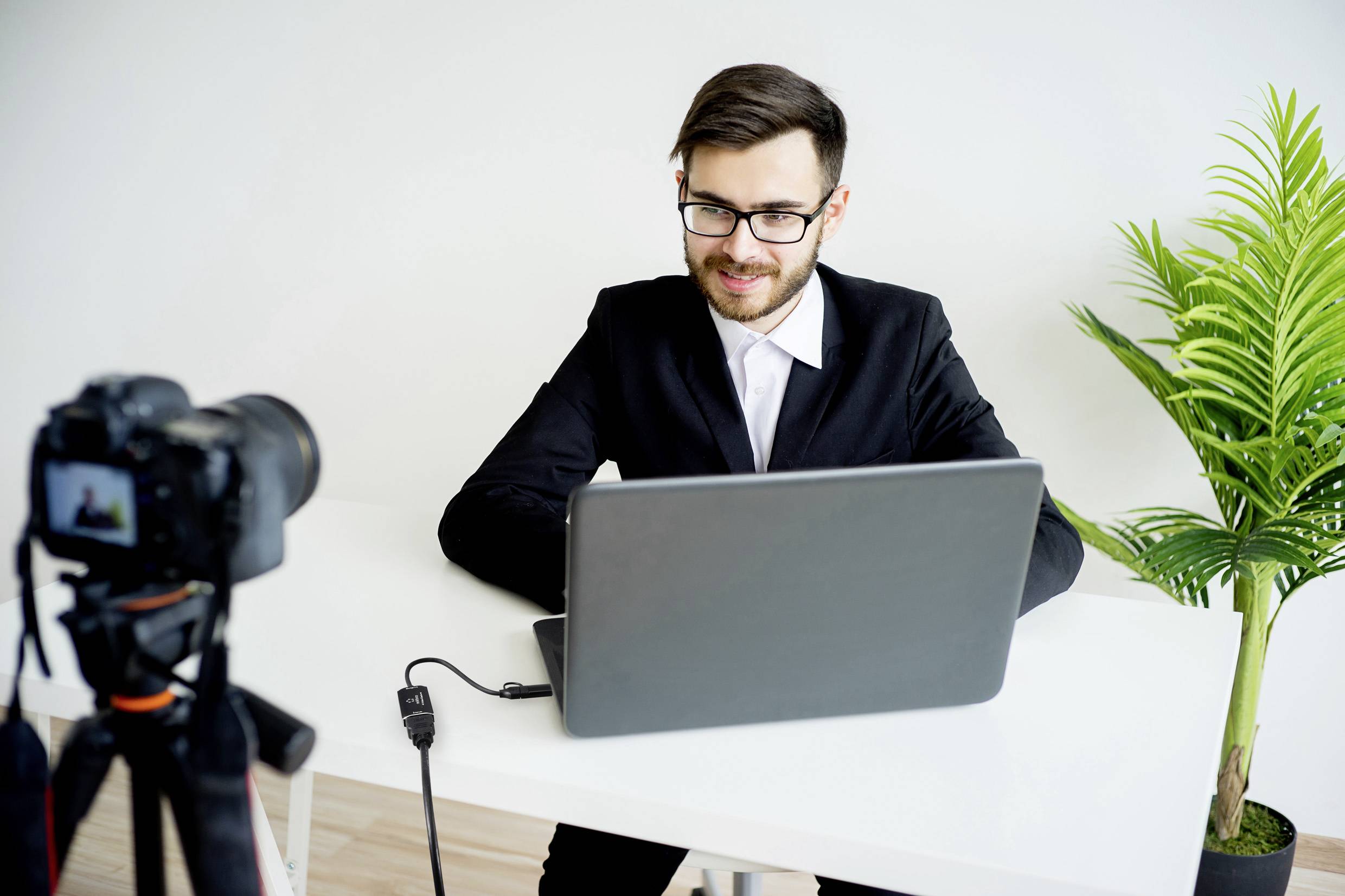 A man in a black suit sits at a table in front of a laptop, looking into a camera. A plant stands to his right.