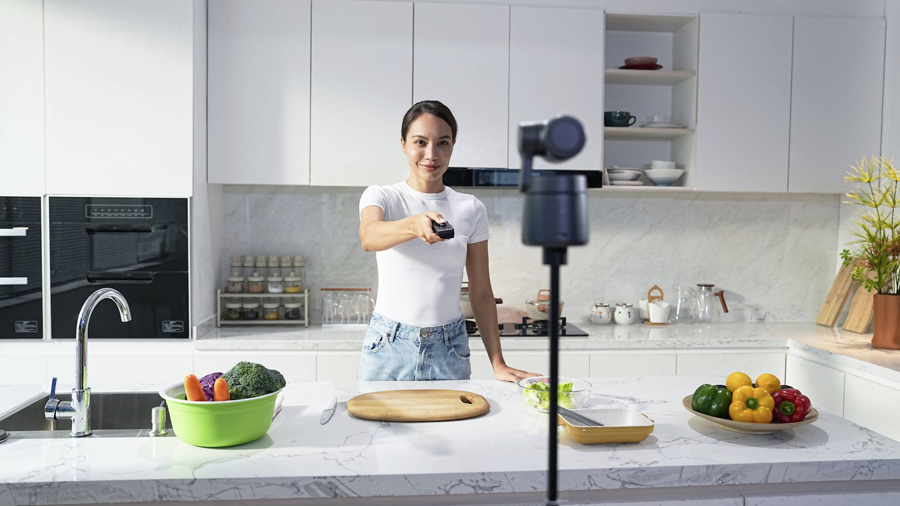 A person is standing in a modern kitchen in front of a camera, holding a remote control and preparing for a recording.