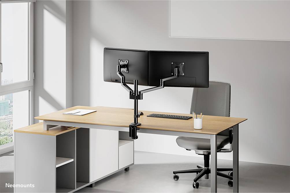 Two monitors mounted on a dual-swing arm bracket on a modern desk in a bright office with a chair.