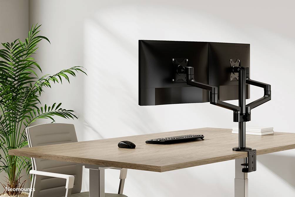Two monitors on adjustable mounting arms on a desk with a keyboard and mouse, next to an office chair and a plant.